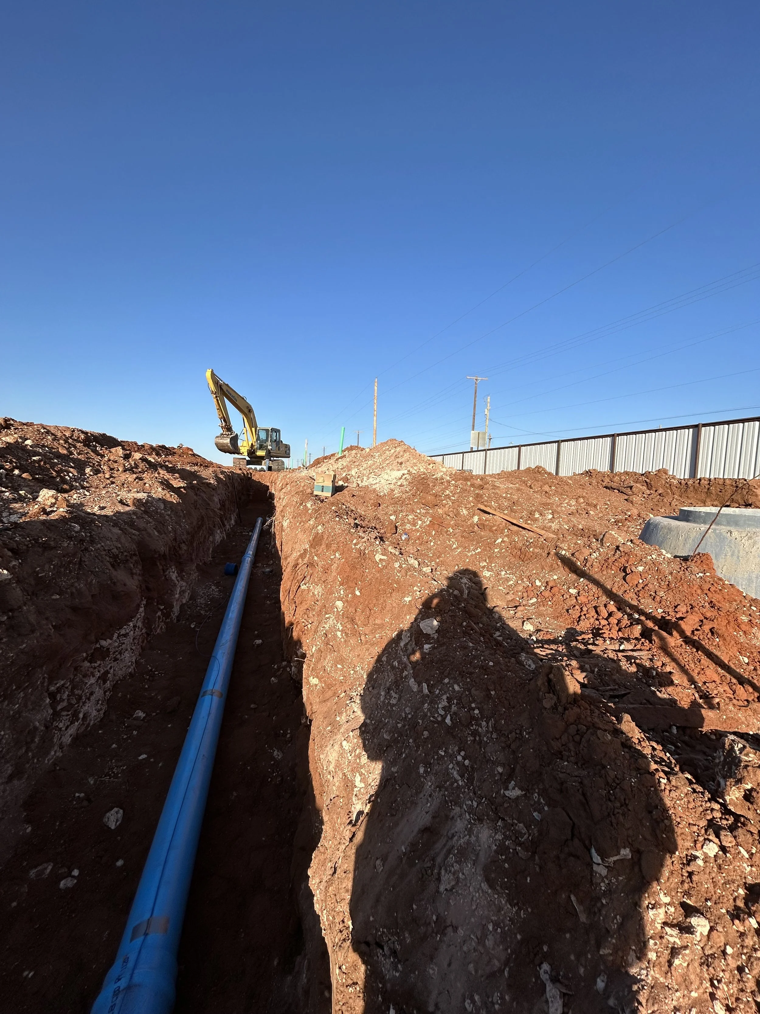 Construction site with excavator and a long blue pipe in a trench, dirt piles, and utility poles under a clear blue sky.