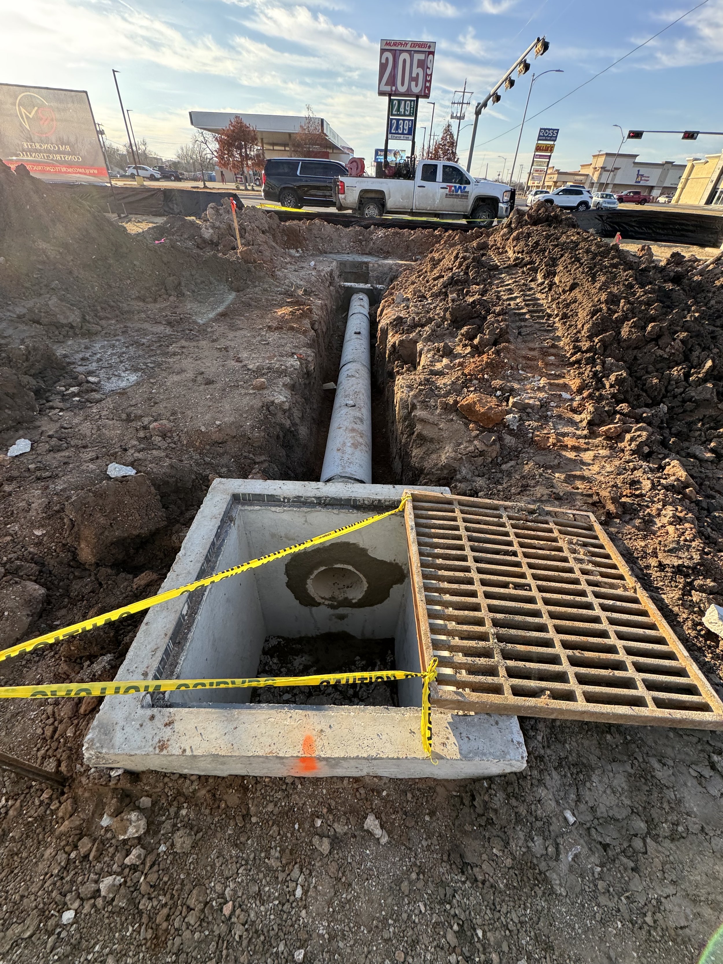 Construction site with an excavated trench containing a large pipe, surrounded by dirt and a yellow caution tape, in an urban area with cars and storefronts in the background.