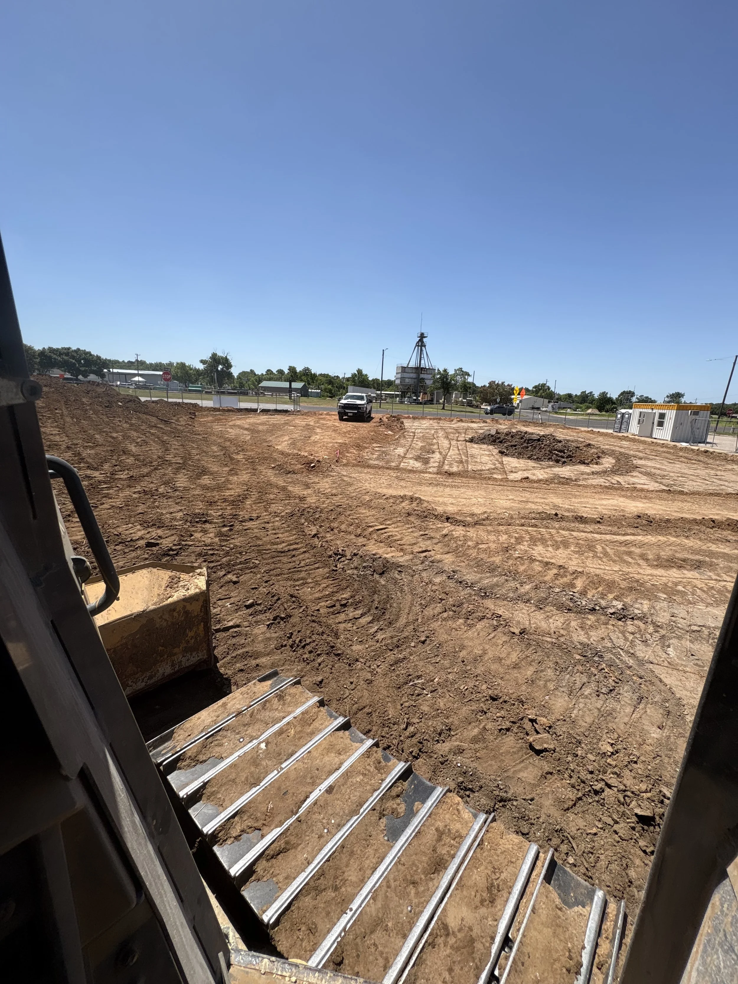 Construction site viewed from inside a vehicle, with metal tracks on the ground and dirt, and a black truck in the distance under a clear blue sky.