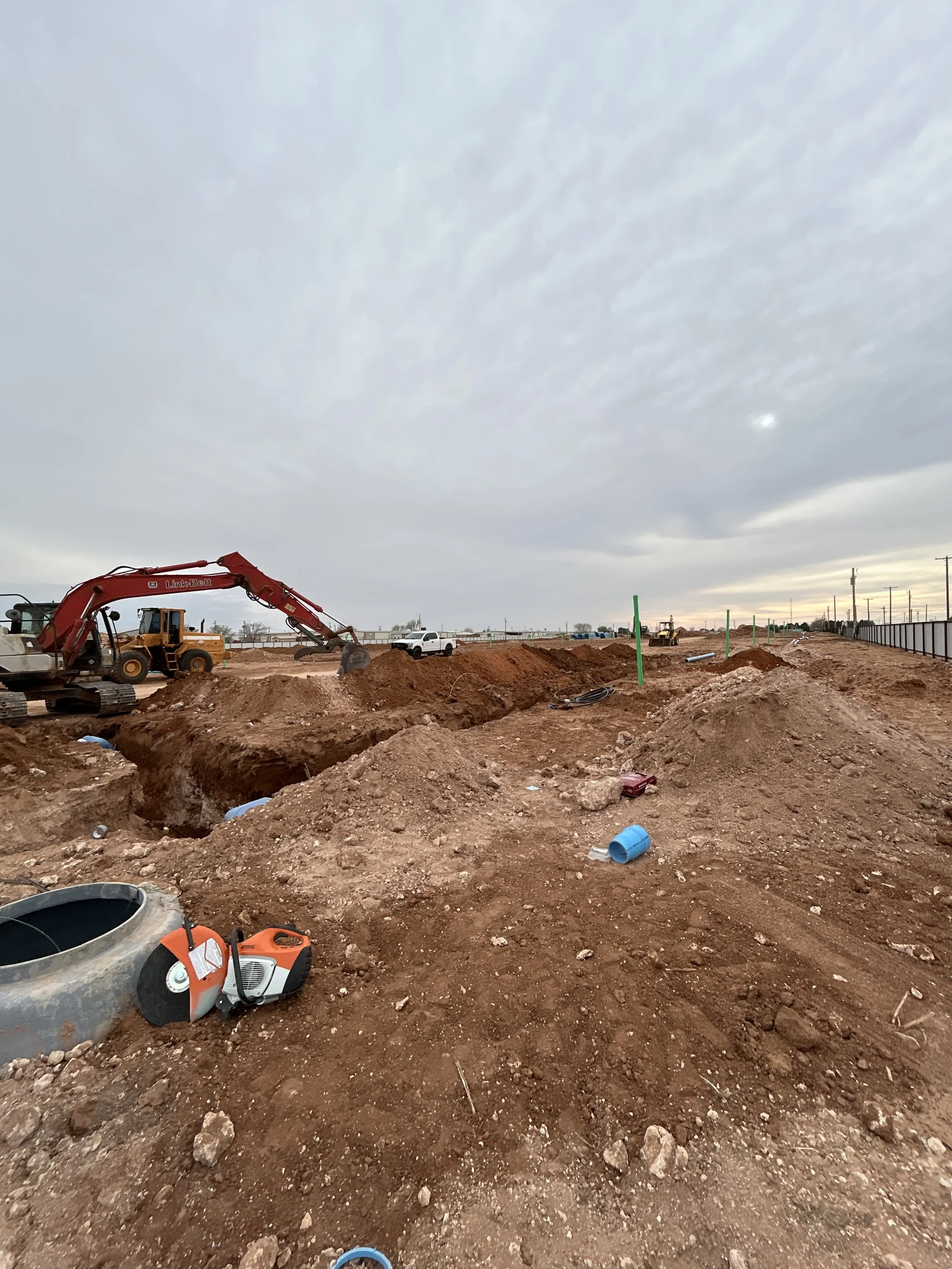 Construction site with excavators and scattered dirt, pipes, and construction tools under cloudy sky.