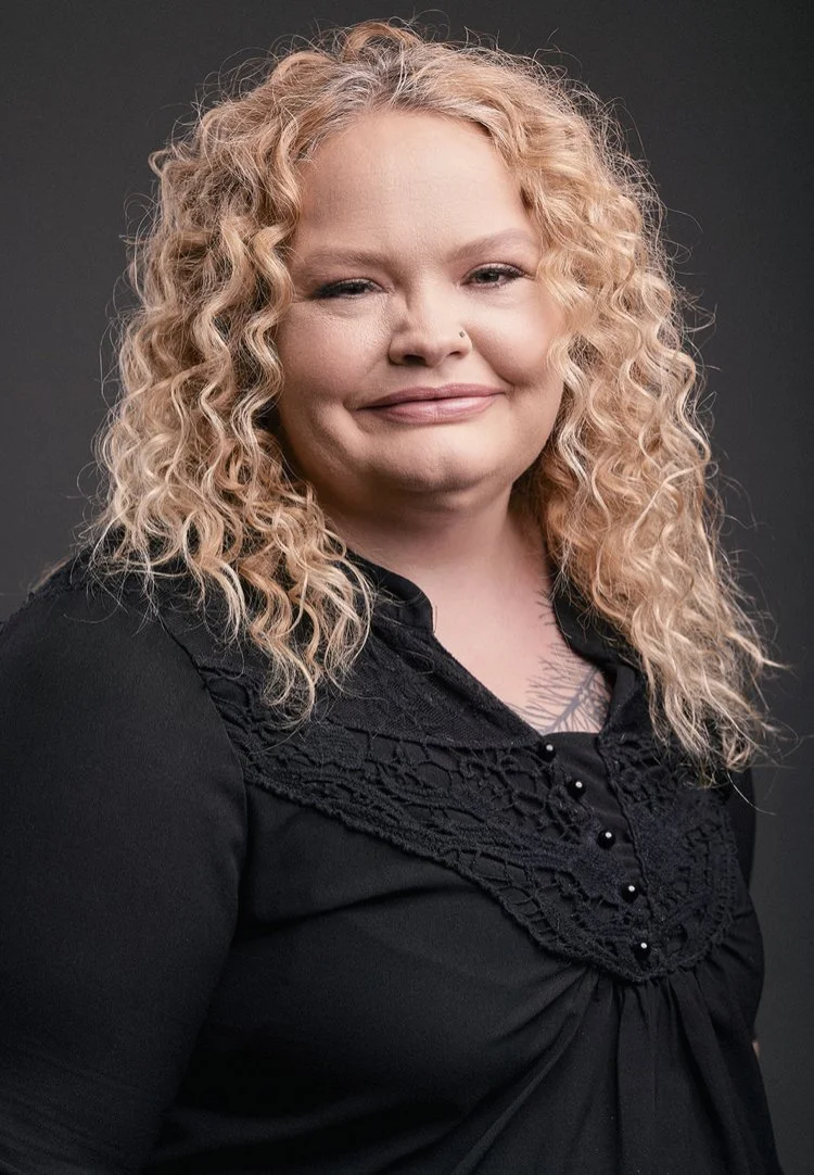 A woman with curly blonde hair wearing a black top with lace and button details, smiling at the camera against a dark background.