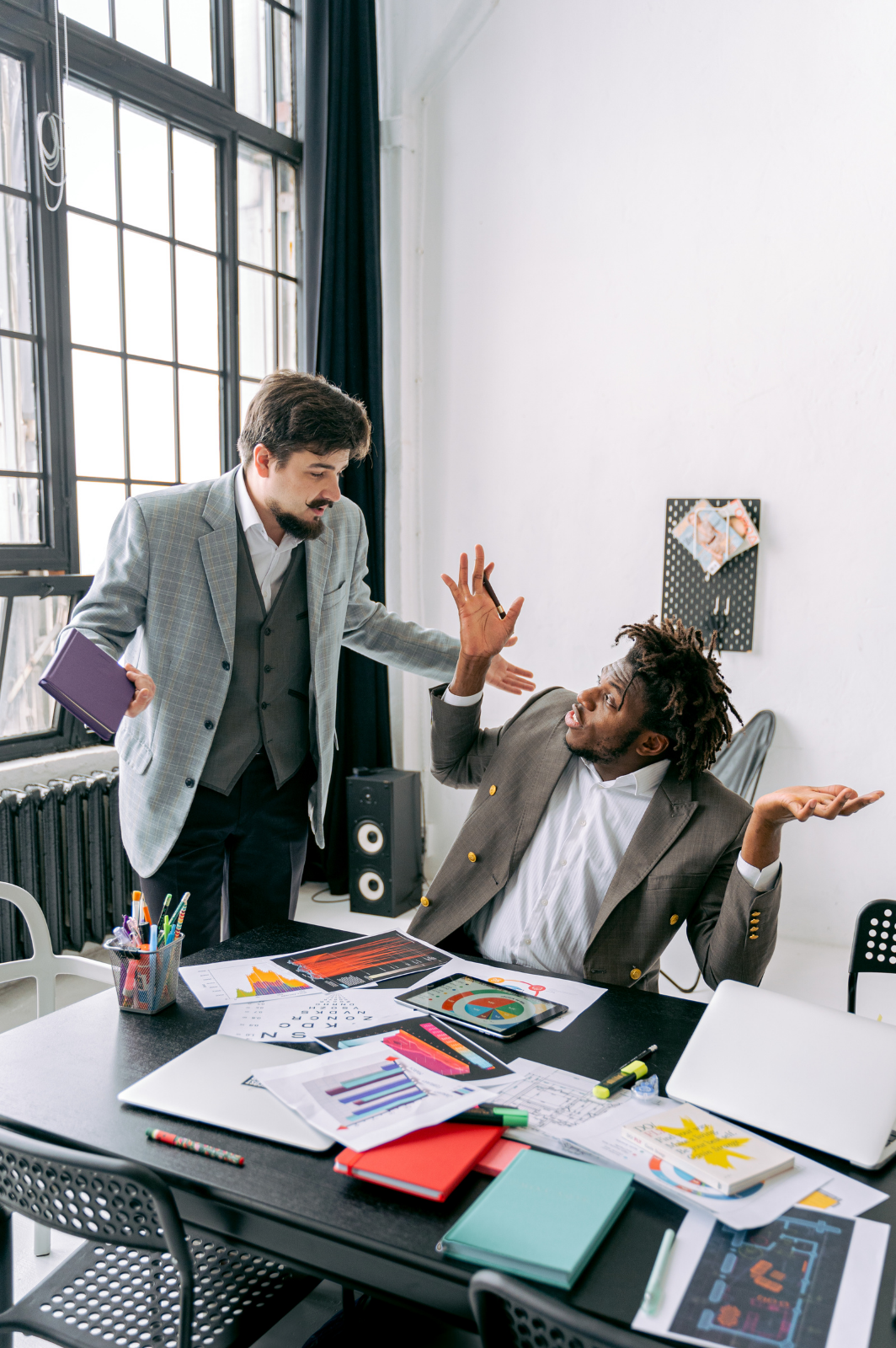 Two men having a heated discussion in an office. One man is standing, holding a purple folder, and the other is sitting, gesturing with his hands. The desk has papers, charts, books, and office supplies.