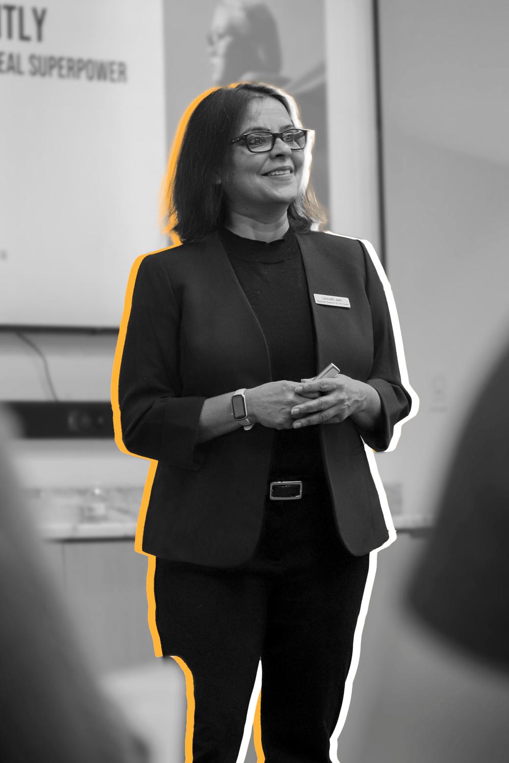 A woman stands smiling, wearing glasses, a dark blazer, and a name tag, in front of a presentation screen with blurred text and images, outlined in yellow.