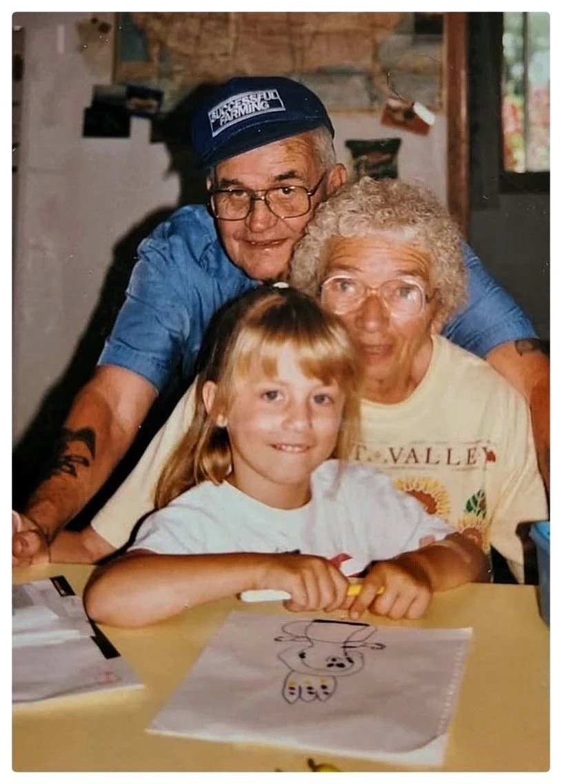 A young girl sitting at a table, smiling, with an alien drawing in front of her. Behind her, two older women and an older man are posing closely together, all smiling.