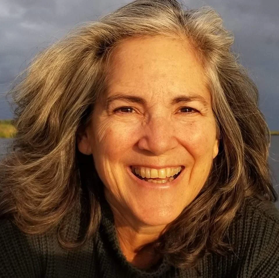 Close-up of a smiling woman with long dark to light brown hair outdoors near water, with a partly cloudy sky in the background.