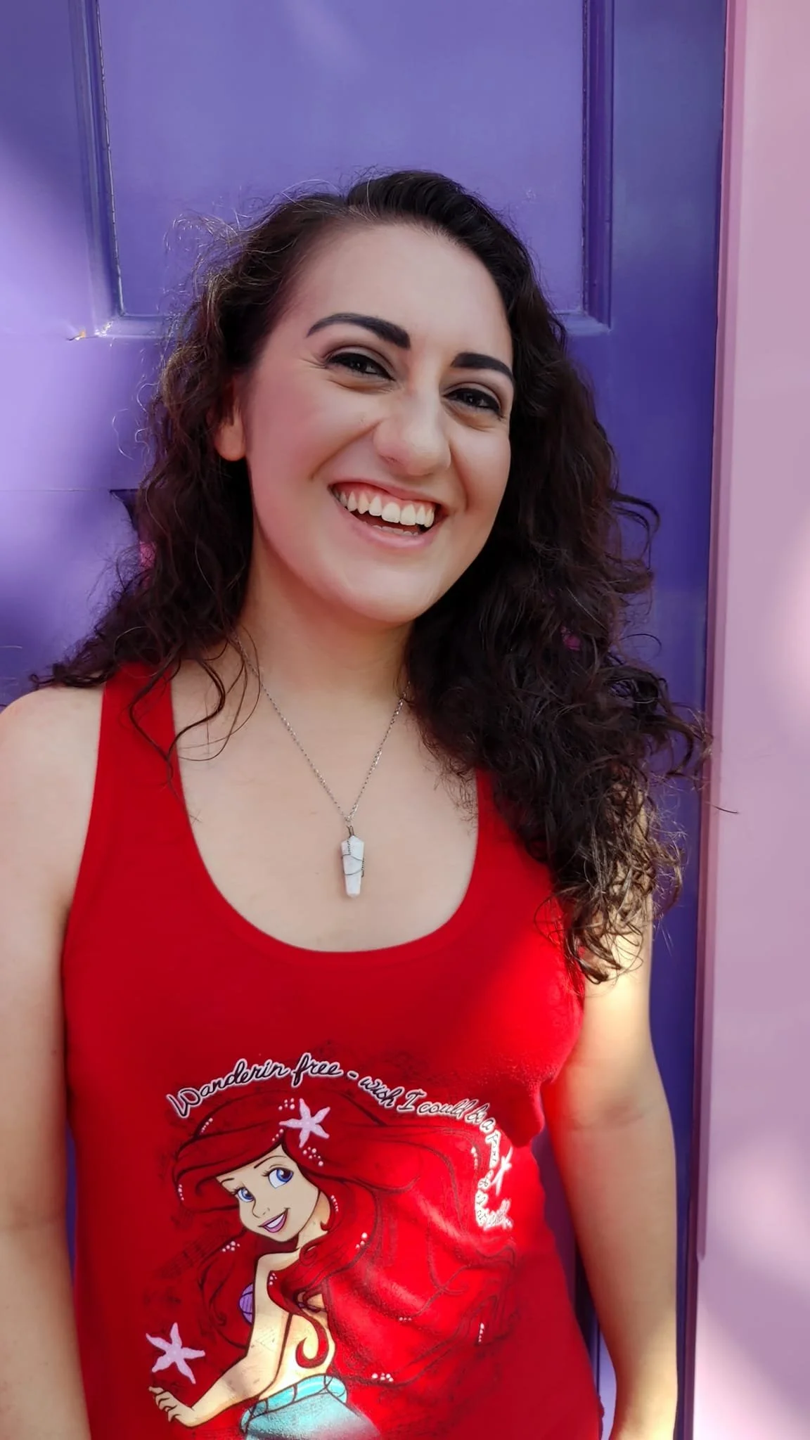A woman with dark curly hair wearing a red tank top featuring a Disney princess design and necklace, standing in front of purple and pink walls, smiling.