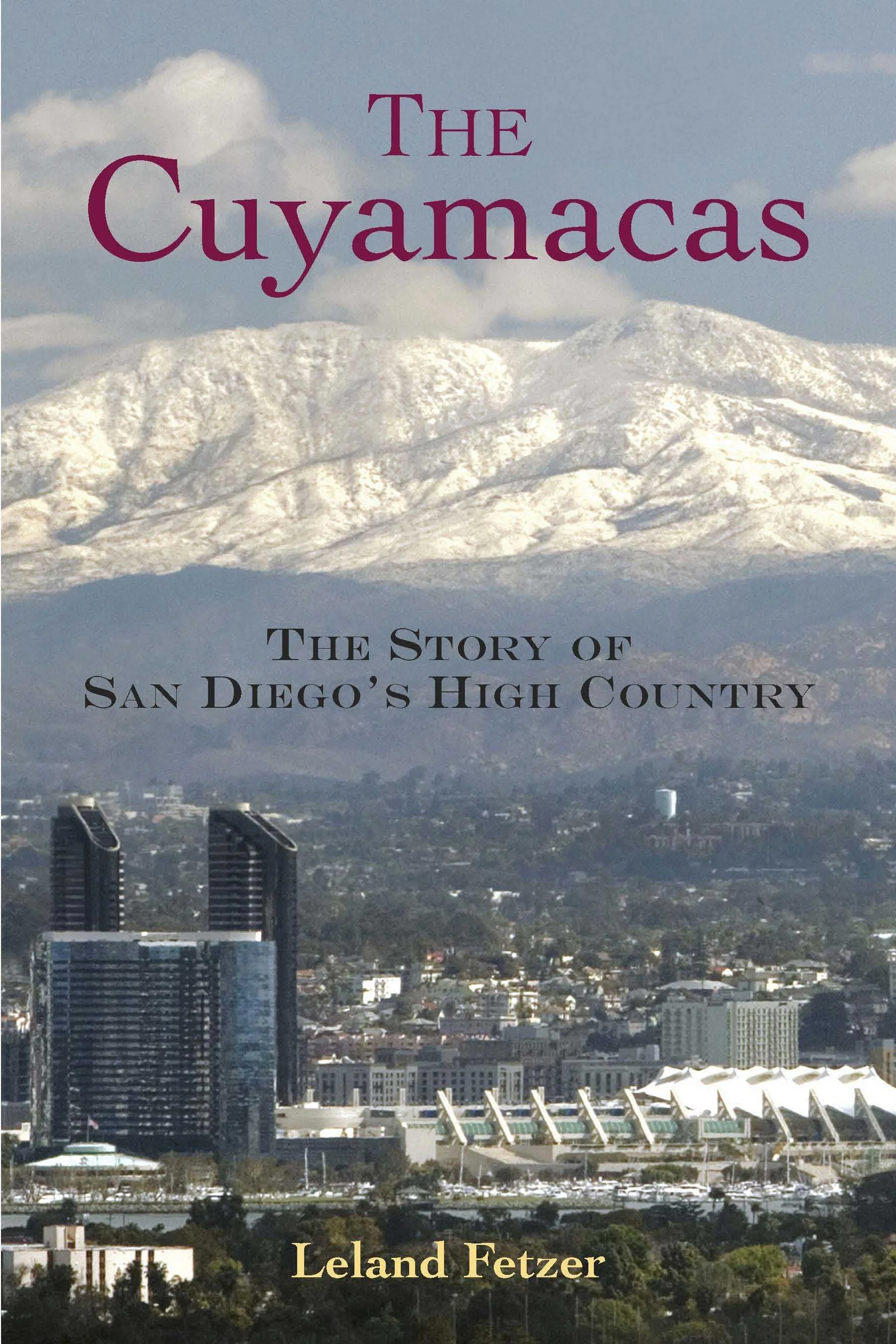Cover of a book titled 'The Cuyamacas' by Leland Fetzer. The subtitle reads 'The Story of San Diego's High Country'. The background features a view of snow-capped mountains and a cityscape with tall buildings and a stadium, under a cloudy sky.