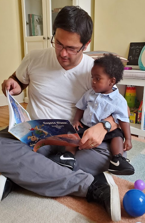 A man with glasses reading a children's book titled 'Cookie Claus' to a young boy sitting on his lap in a colorful playroom.