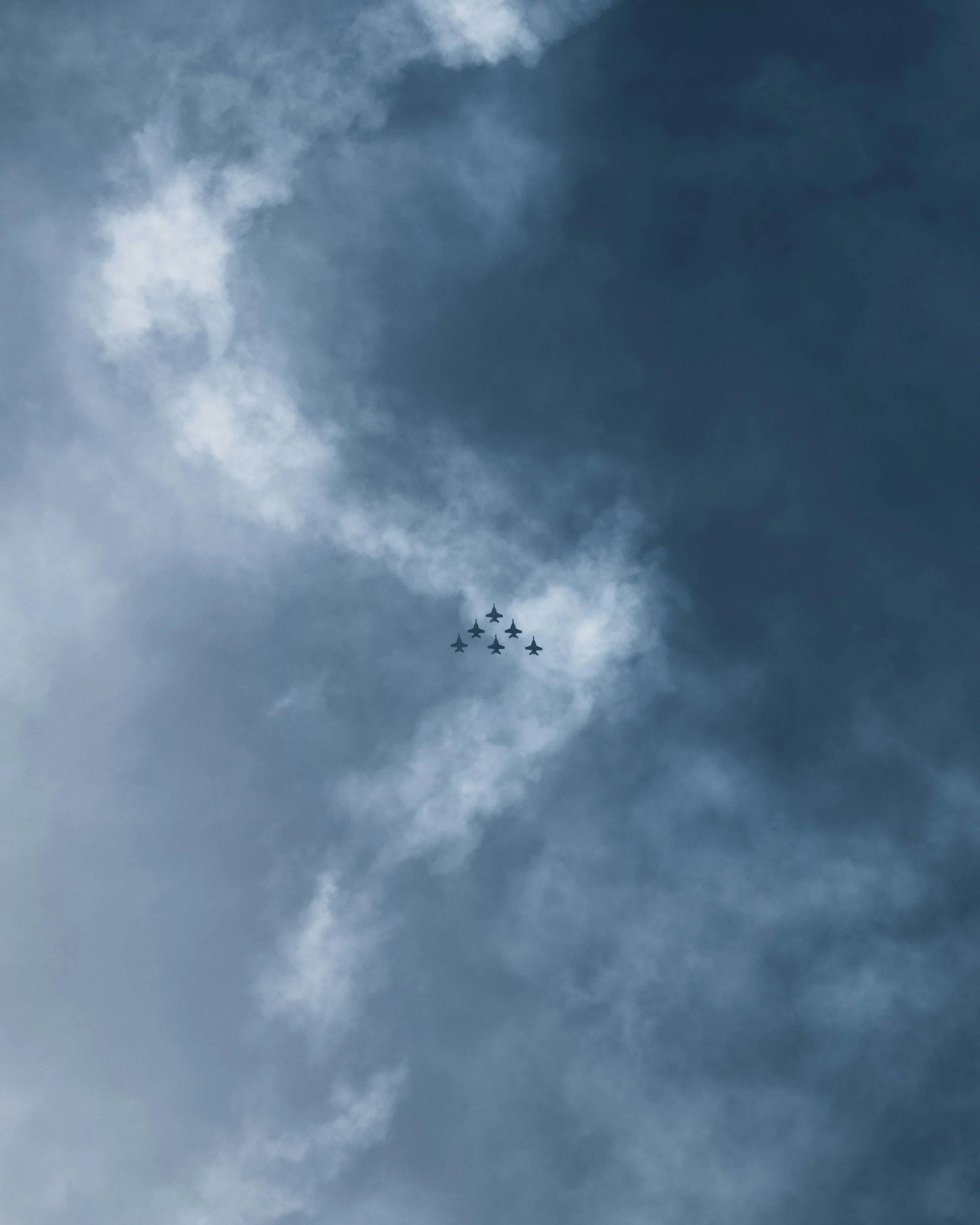 Formation of seven military fighter jets flying in a tight V-shape pattern with trails of afterburner exhaust against a blue sky with clouds.