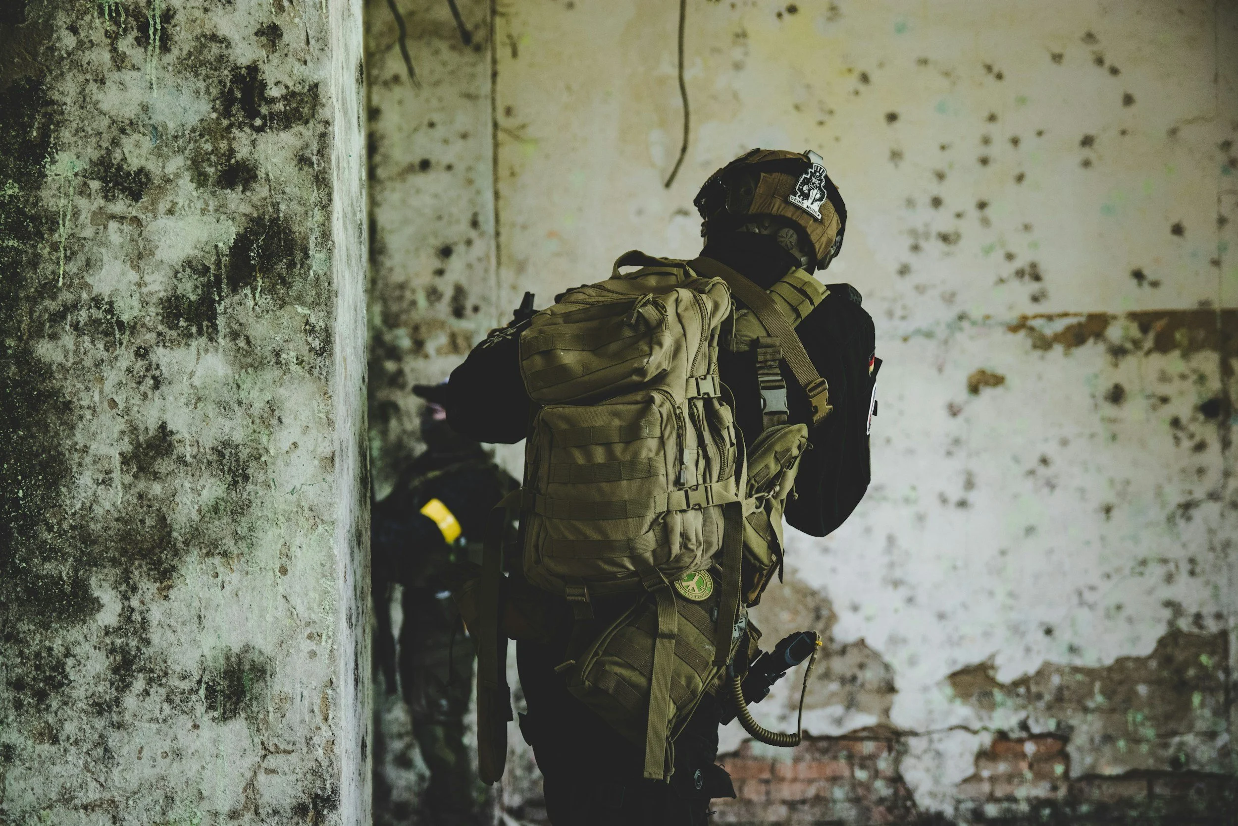 Person in tactical gear wearing a helmet and backpack, standing in a deteriorated, graffiti-covered urban setting.