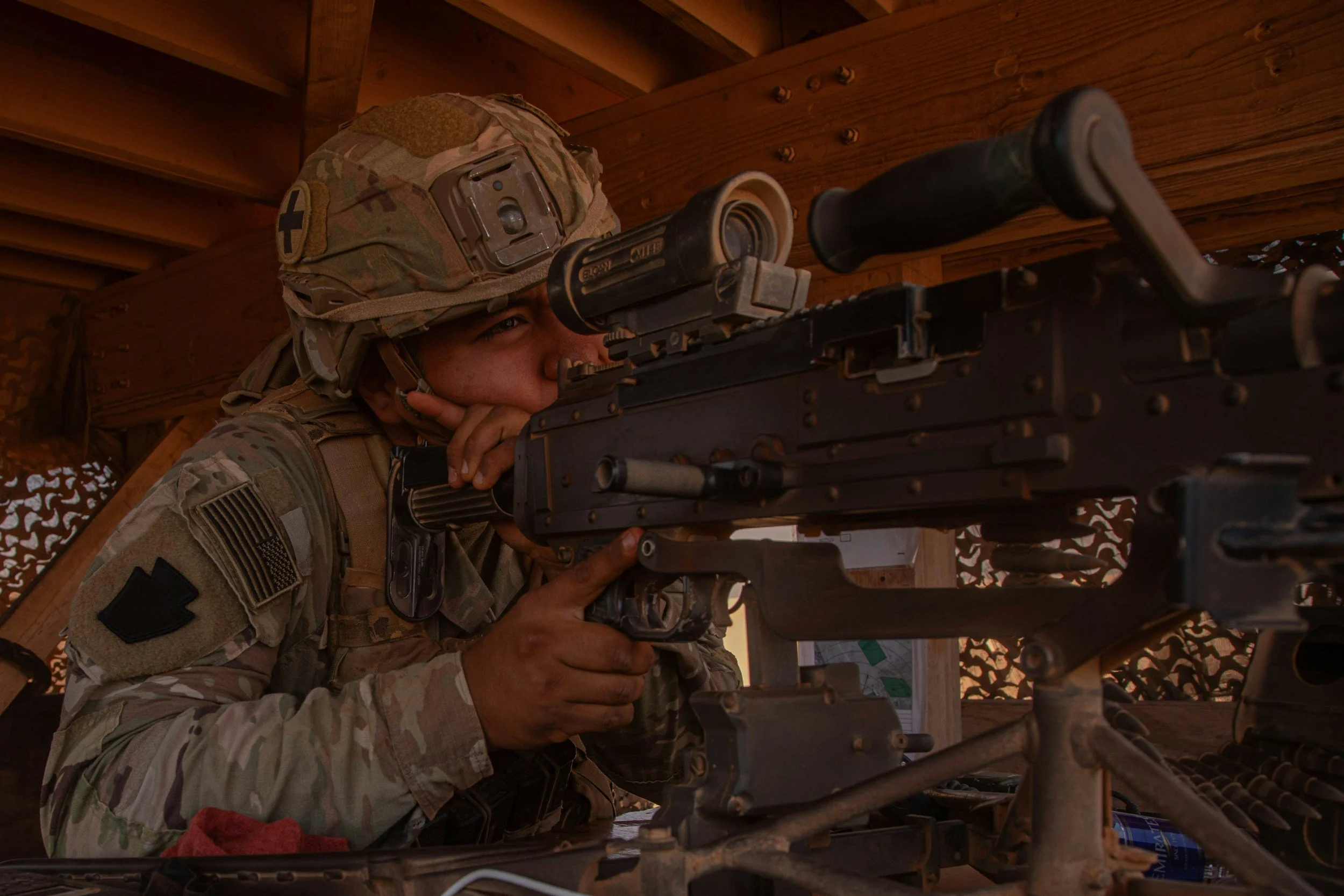 A soldier in camouflage uniform and helmet aiming a sniper rifle while resting her head on her hand in a wooden shelter.