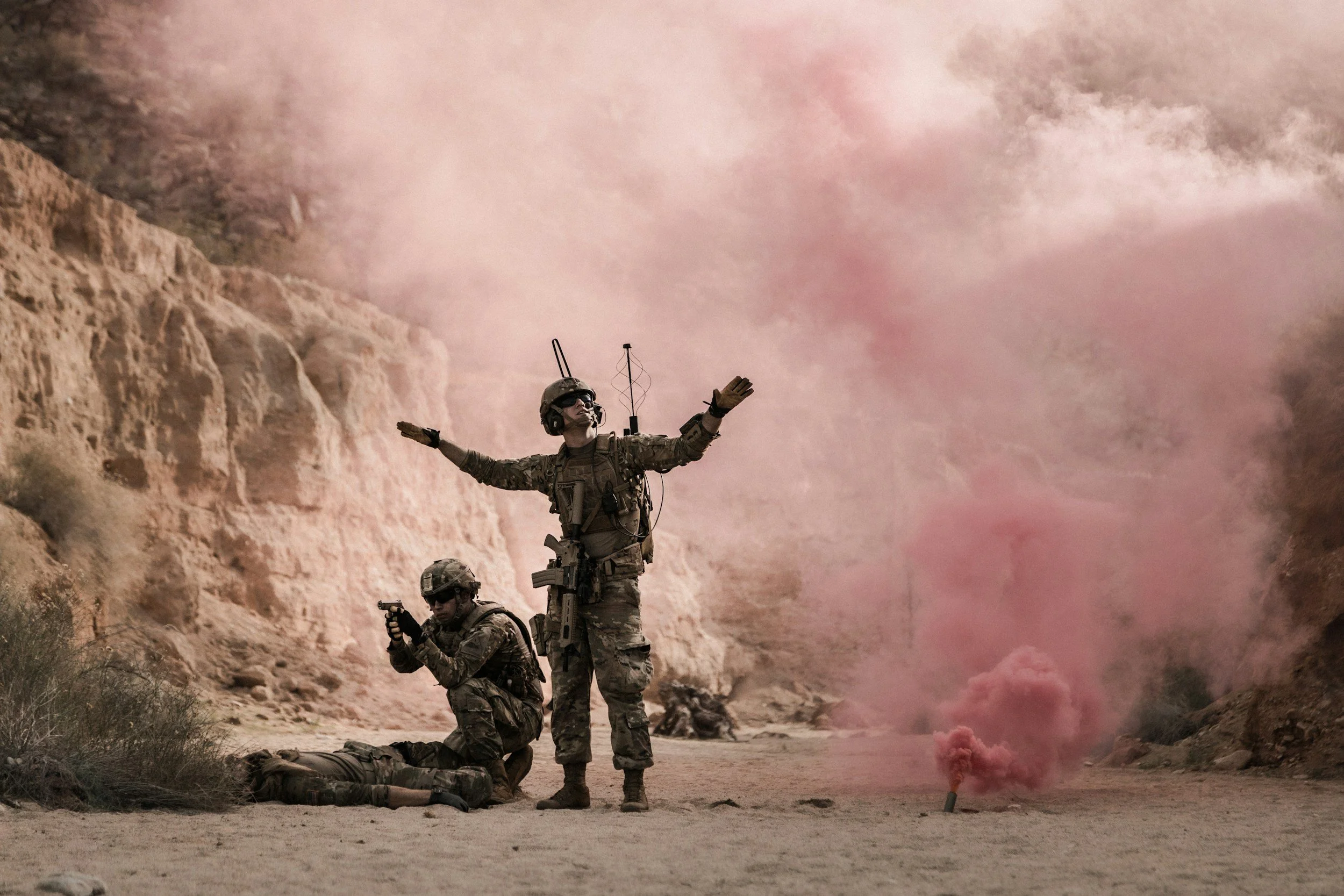 Two soldiers in camouflage uniforms in a desert landscape with rocky cliffs, one standing with arms outstretched, another kneeling aiming a gun, and the third lying on the ground, surrounded by pink smoke.