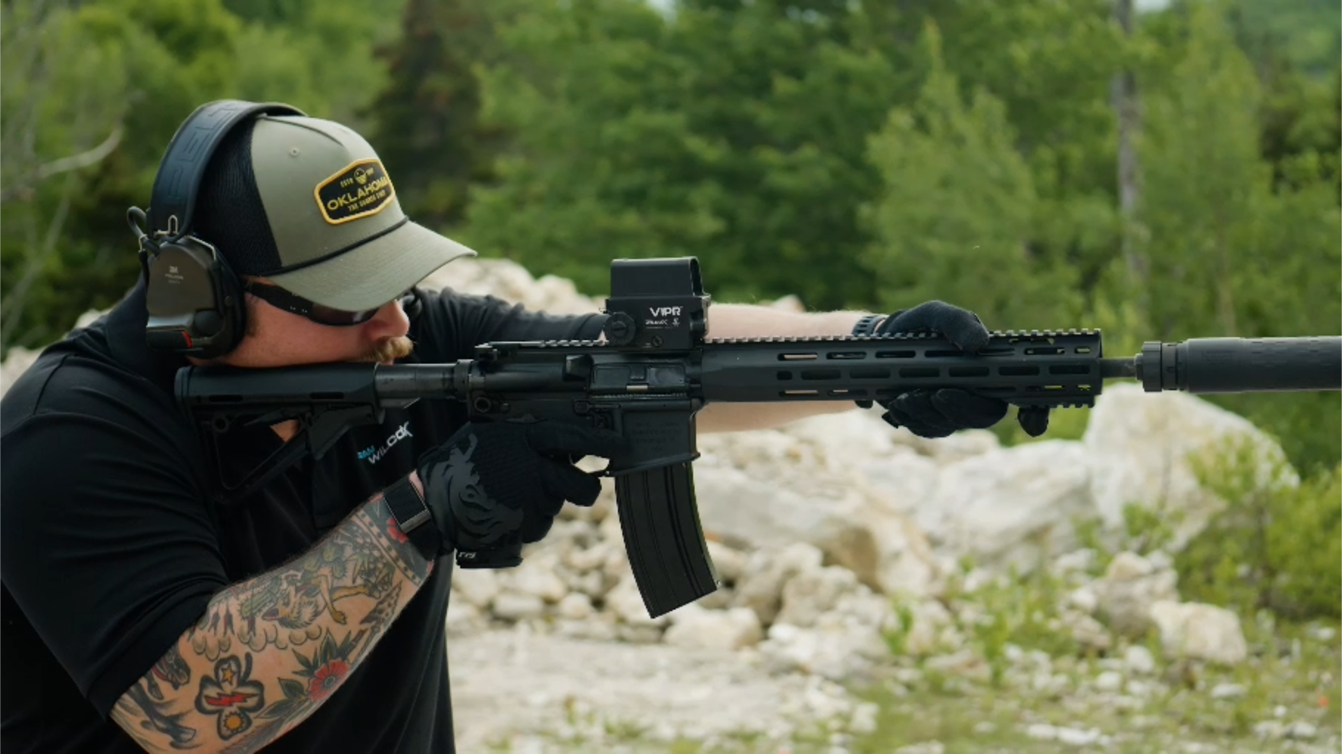 A man with colorful tattoos on his forearm, wearing a gray cap, black shirt, and protective glasses, aiming a rifle with a scope in an outdoor setting with green trees and rocks.