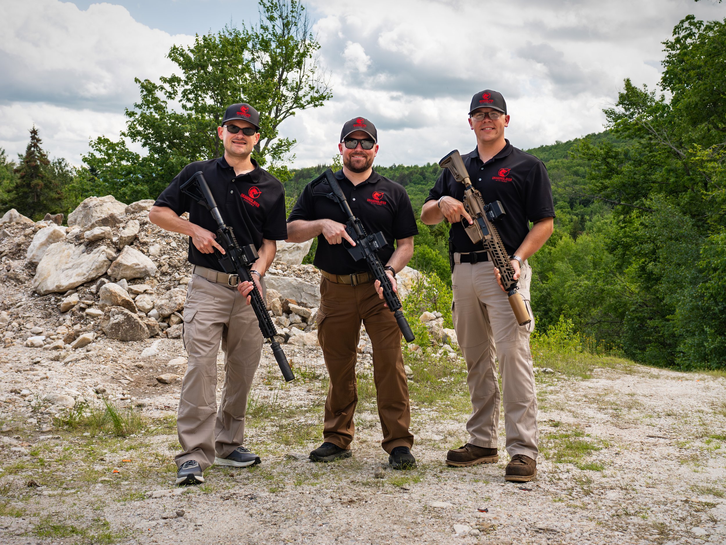 The Dragonslayer System team standing outdoors on rocky terrain holding rifles, dressed in black shirts with red Dragonslayer logos, and wearing caps. Trees and hills are visible in the background under a partly cloudy sky.