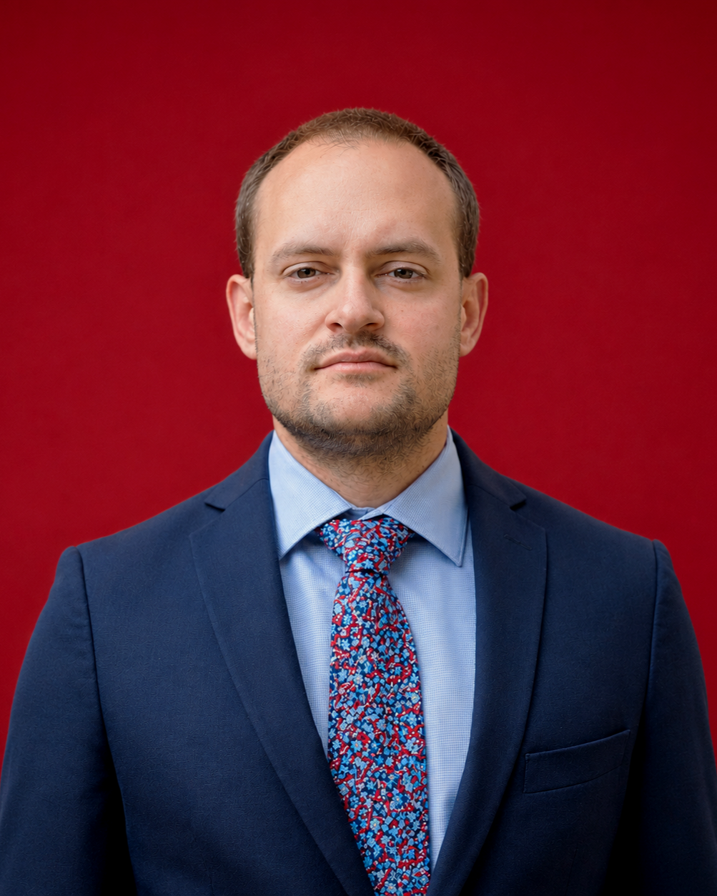 A man in a blue suit, light blue shirt, and floral tie standing against a red background.