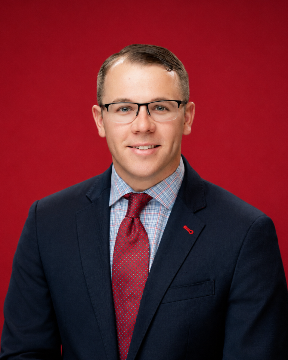A man wearing glasses, a navy blue blazer, a checked shirt, and a red tie, standing against a red background.