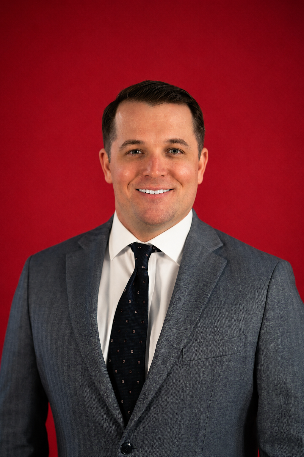 Nick Culbert in a gray suit, white shirt, and black tie smiling against a red background.