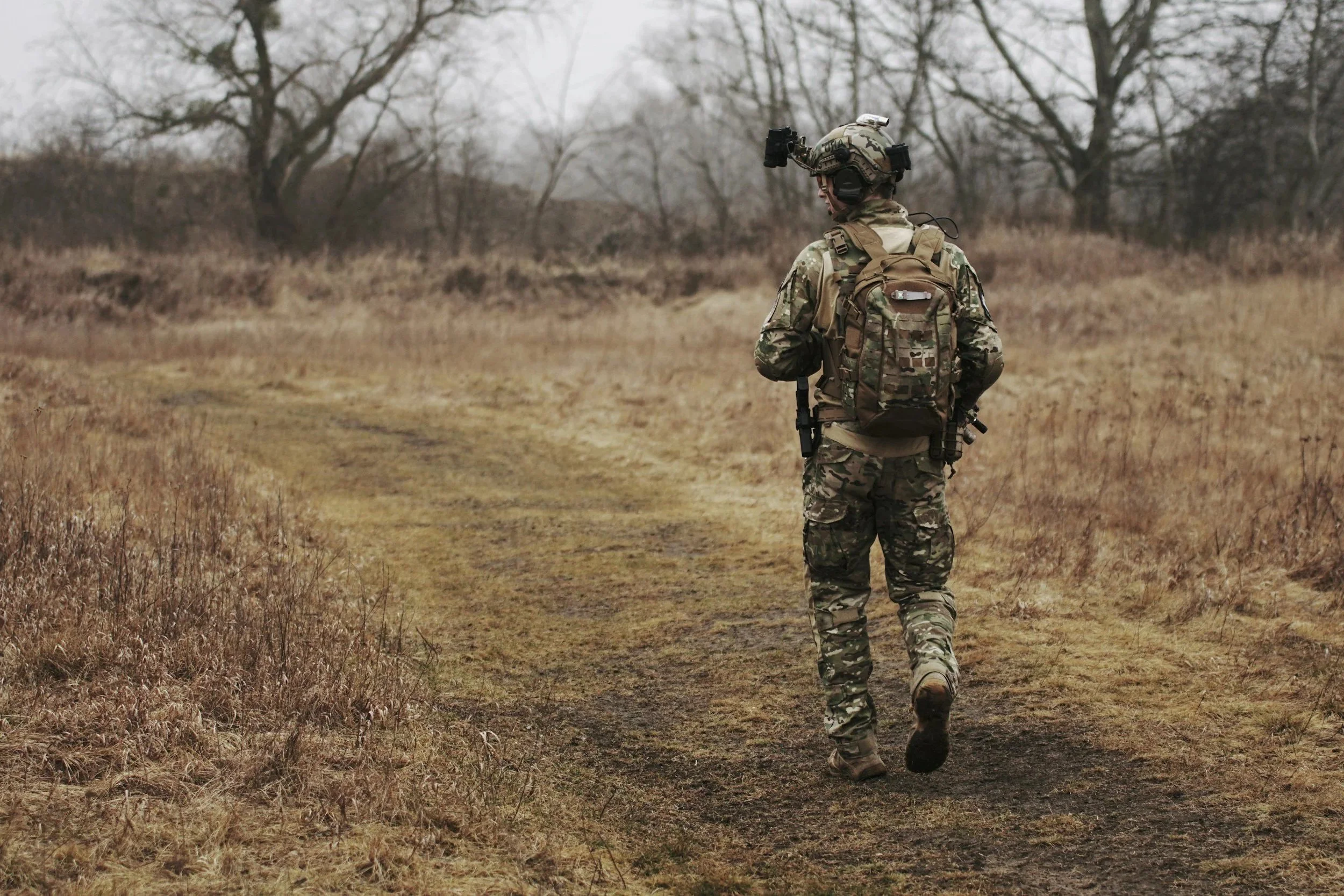 A soldier in camouflage uniform walking along a dirt path through a dry, wooded area on a cloudy day, carrying a backpack and wearing a helmet with mounted equipment.