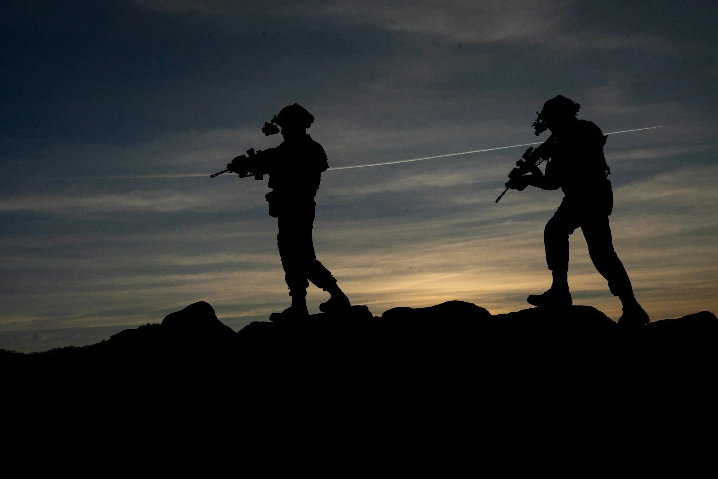 Silhouettes of two soldiers wearing helmets and tactical gear walking on rocky terrain during sunset or sunrise.