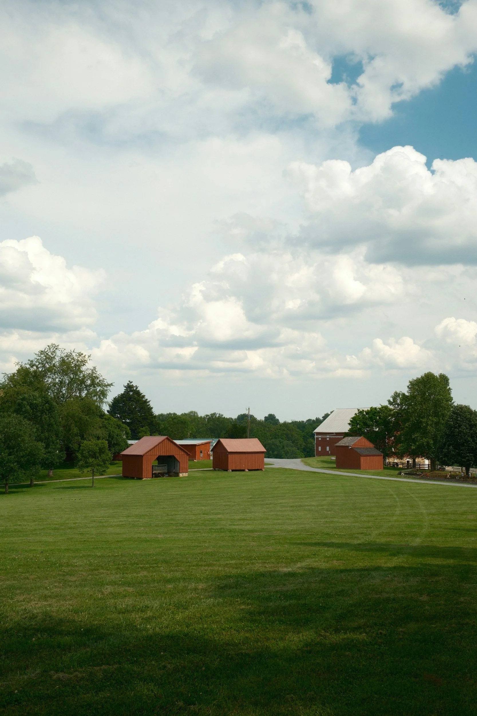 A rural landscape with red barns and farm buildings surrounded by green grass, trees, and a winding road, under a partly cloudy sky.