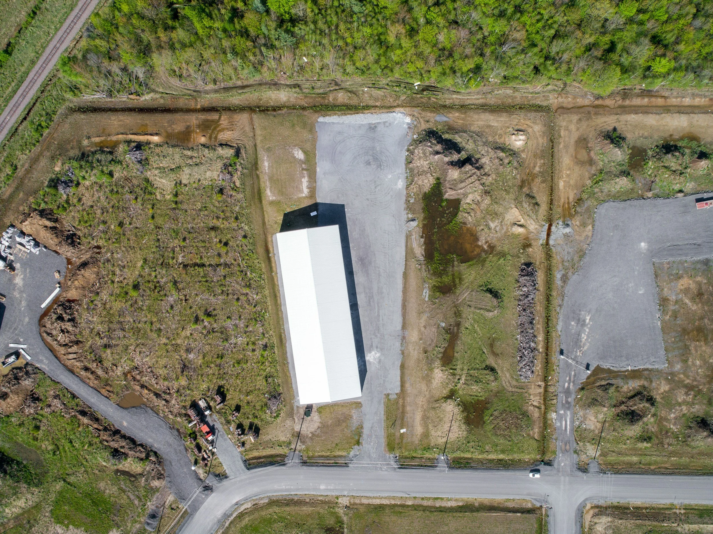 Aerial view of a construction site and a warehouse building surrounded by greenery, roads, and a parking lot.