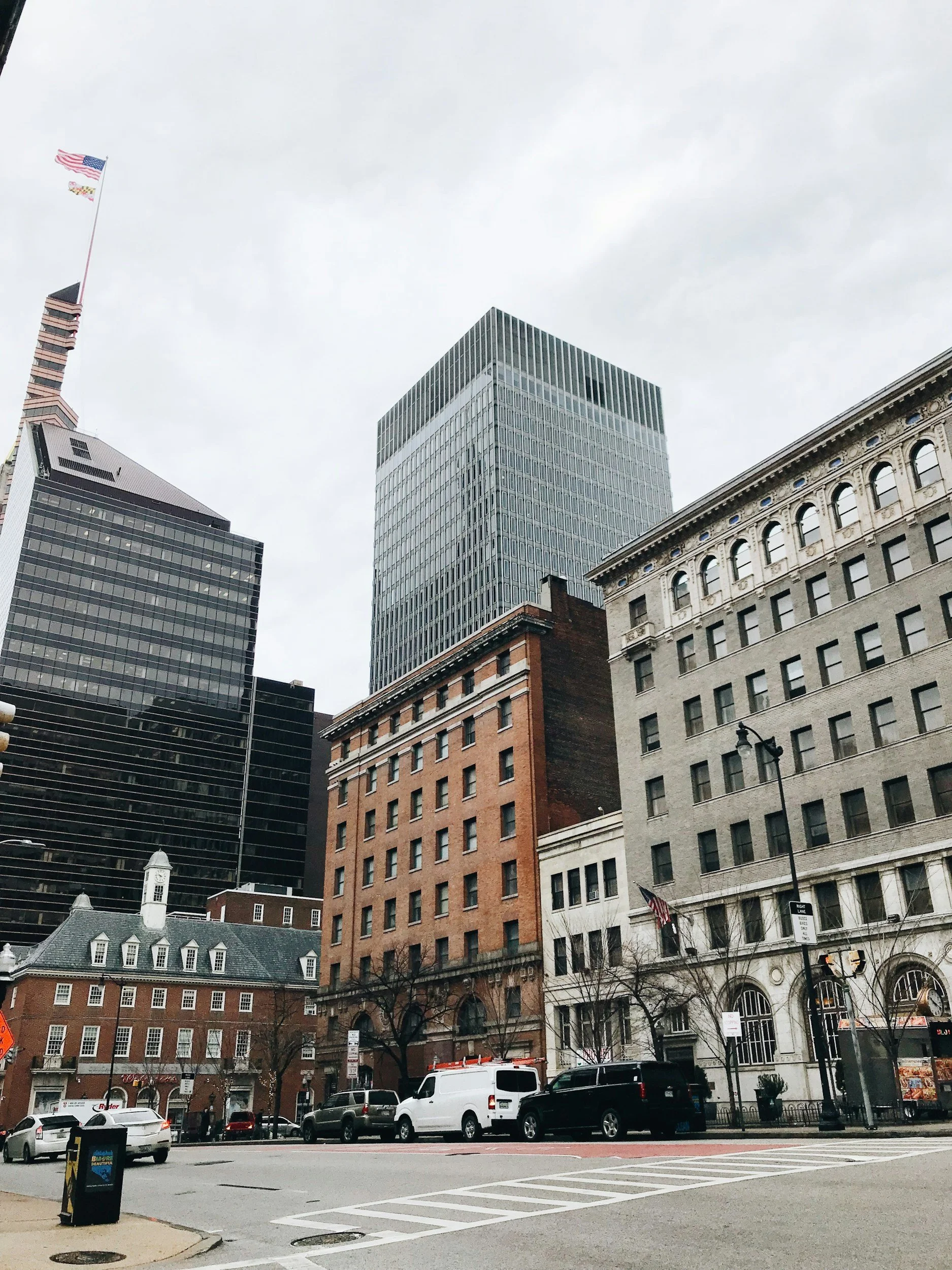 City street scene with tall office buildings, parked cars, and storefronts, overcast sky.