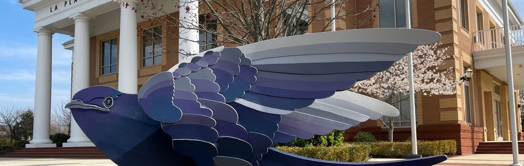 Blue and purple painted sculpture of a bird with wings spread, situated outside a brick building with white columns and cherry blossom trees.