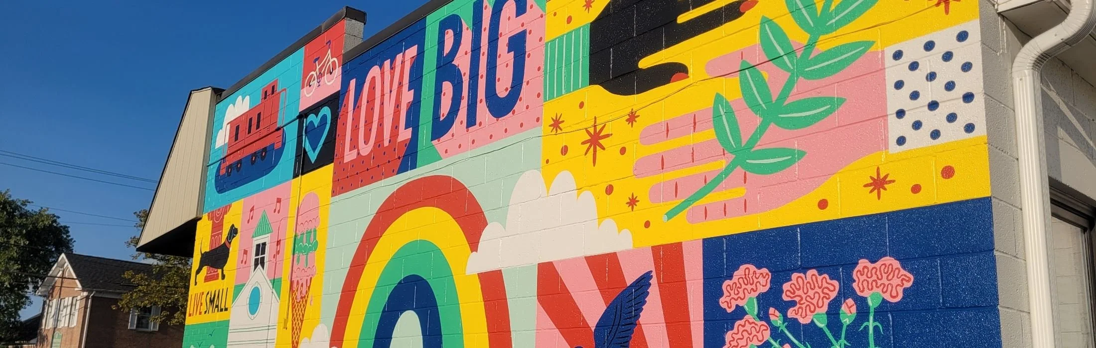 Colorful mural on a building featuring a rainbow, flowers, a rainbow, a dog, a pie, a rainbow, and text that says "Luv It Up" and "Live Small" against a clear blue sky.