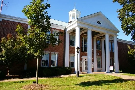 A brick building with white columns and a small white cupola on top, surrounded by trees and a well-maintained lawn with a lamp post in front.