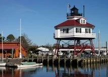 A white lighthouse with a red roof near a body of water, with boats docked along the pier.