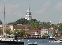 View of a lighthouse on a hill overlooking a waterfront with boats docked in the foreground and buildings along the shoreline.