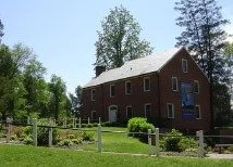 A brick house with a gabled roof, surrounded by green trees and a wooden fence.