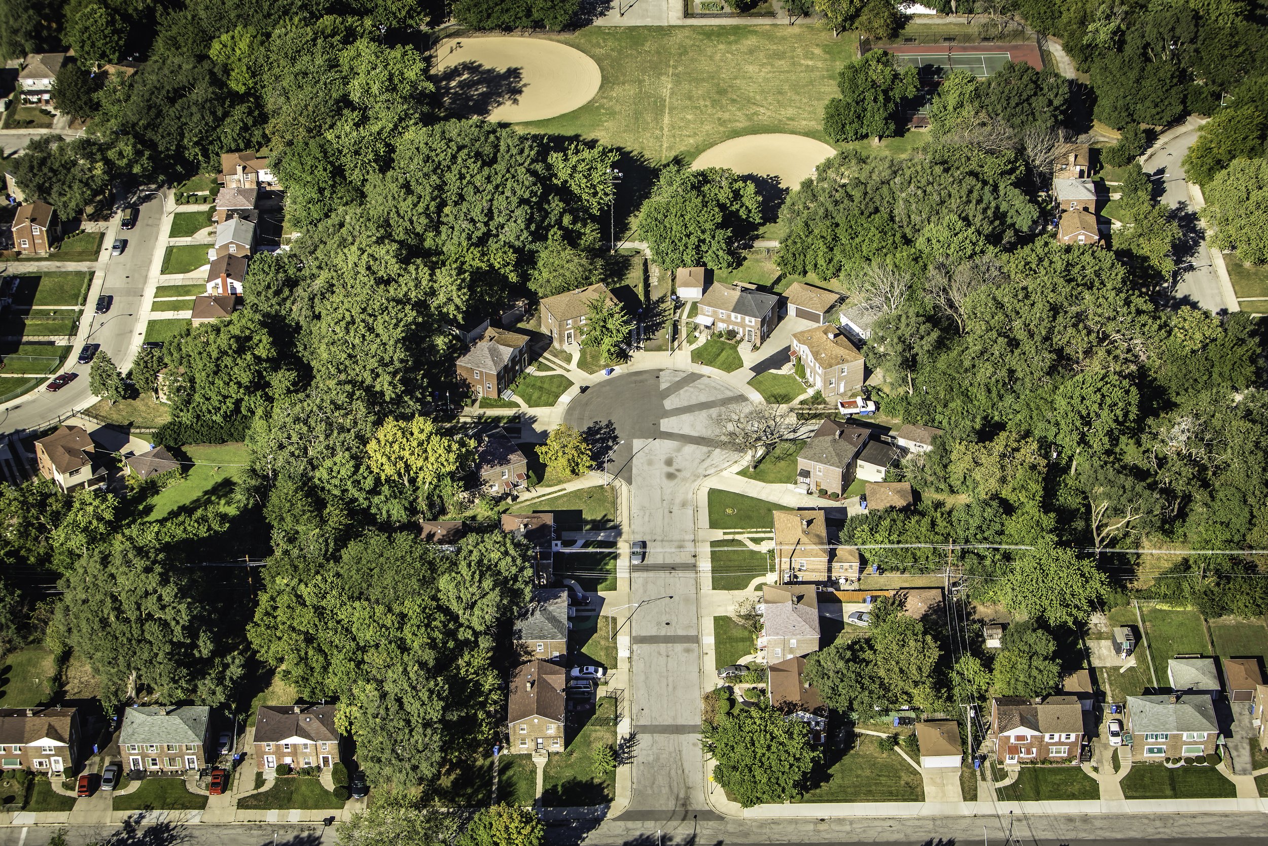 An aerial view of a residential neighborhood with houses, trees, and a green park containing two baseball/softball fields and a smaller sports court.