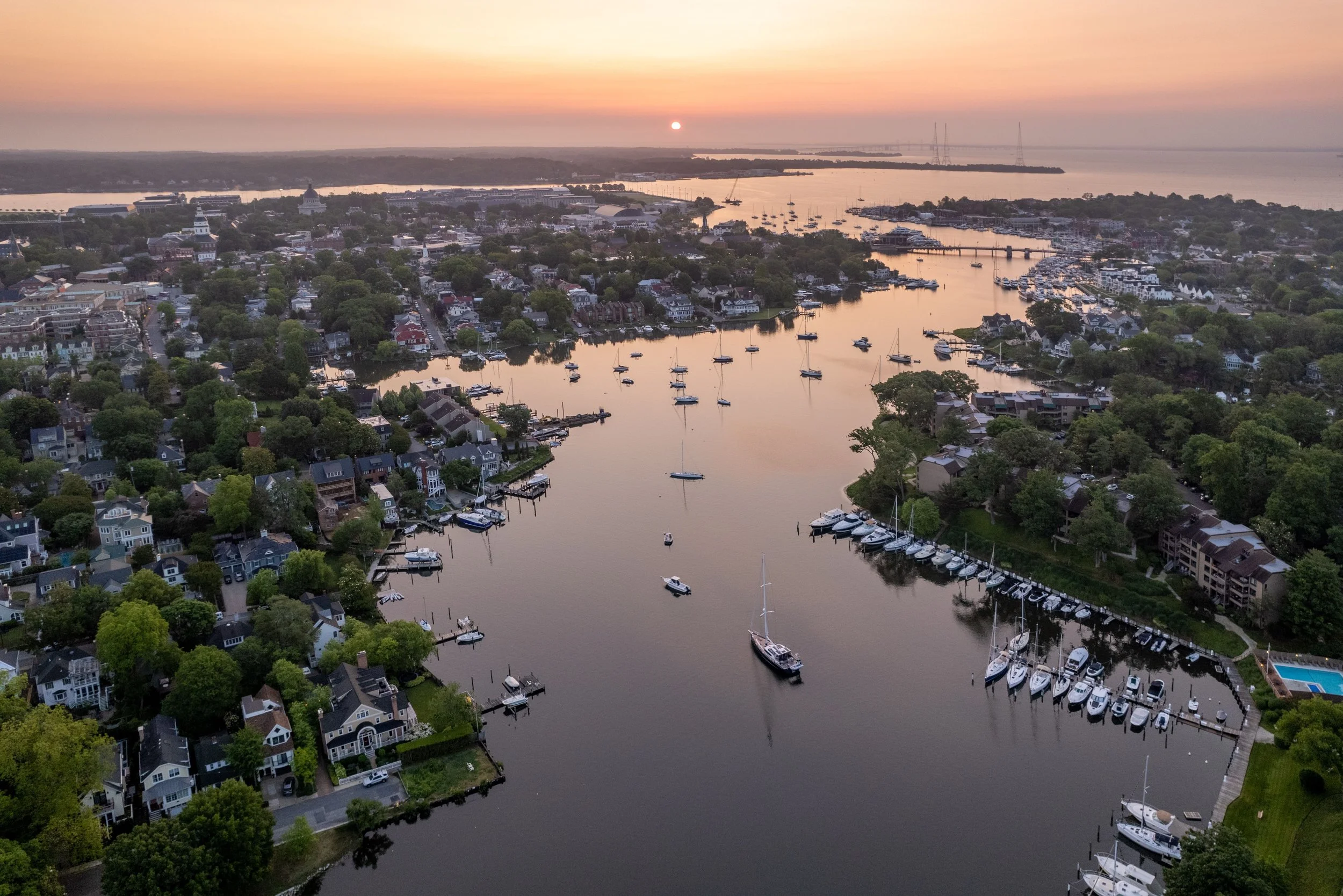 Aerial view of a marina with boats, surrounded by a residential area with trees, under a sunset sky over a body of water.