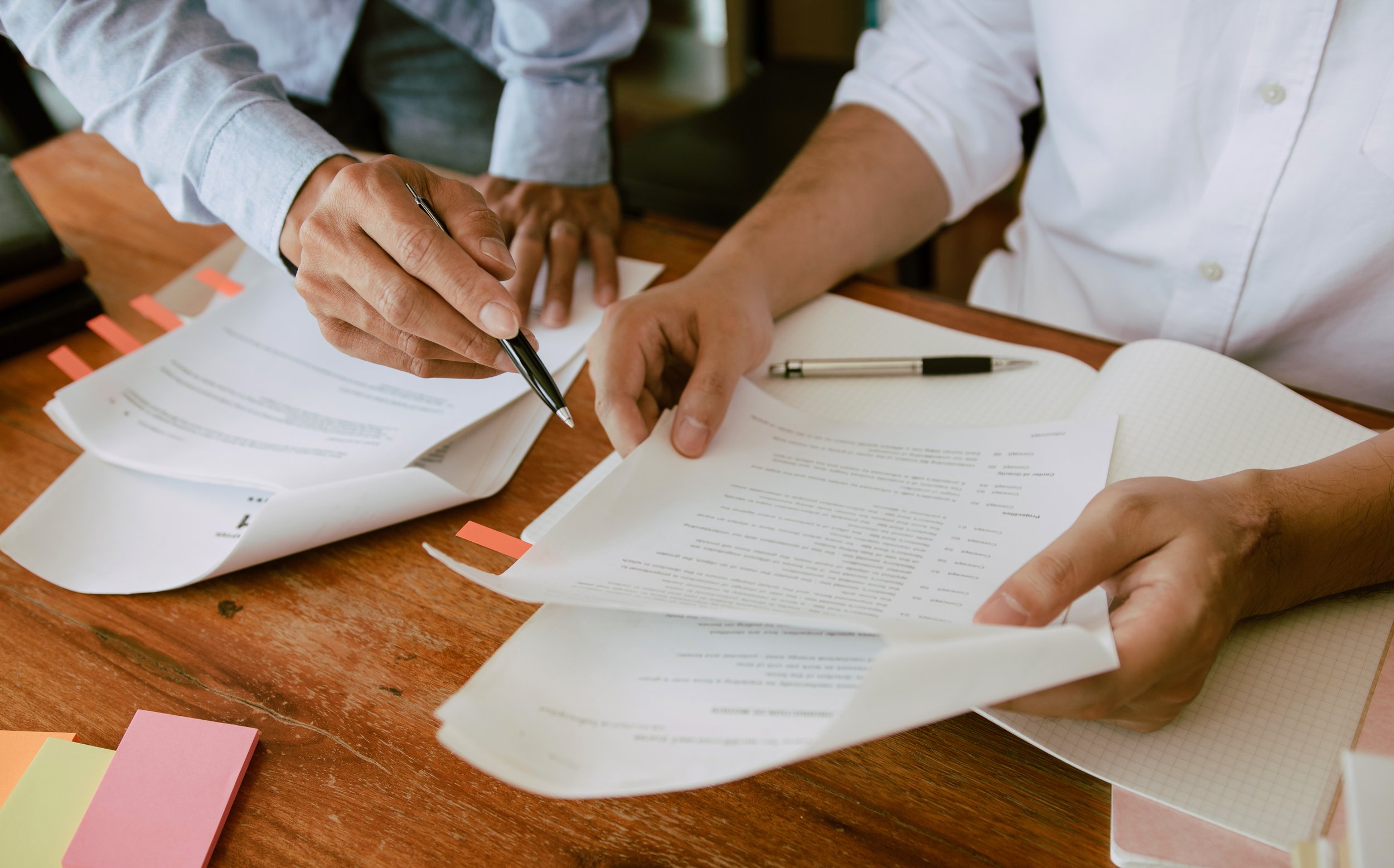 Two individuals reviewing documents at a wooden table with scattered sticky notes.