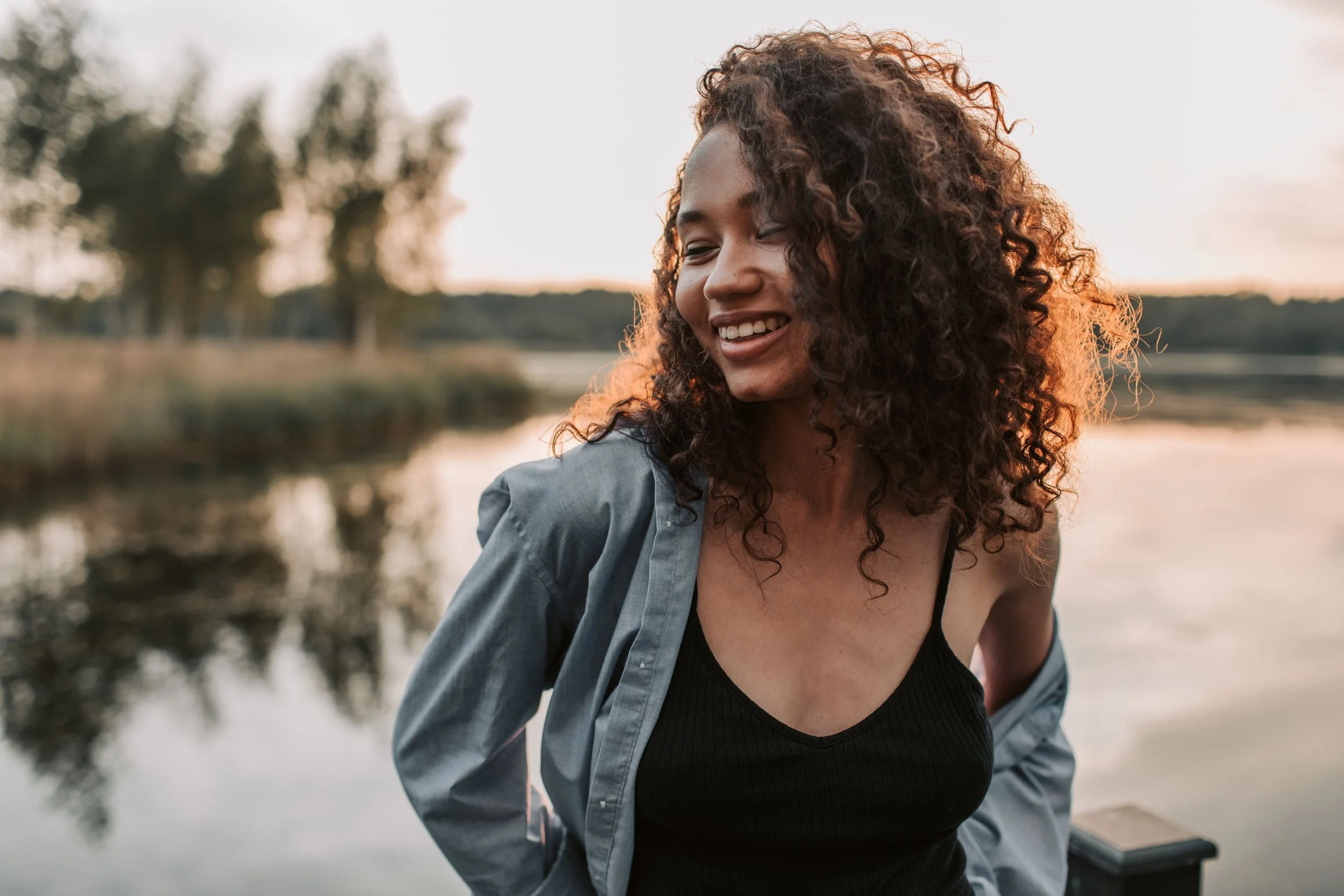 A woman with curly hair smiling while outdoors near a body of water at sunset.