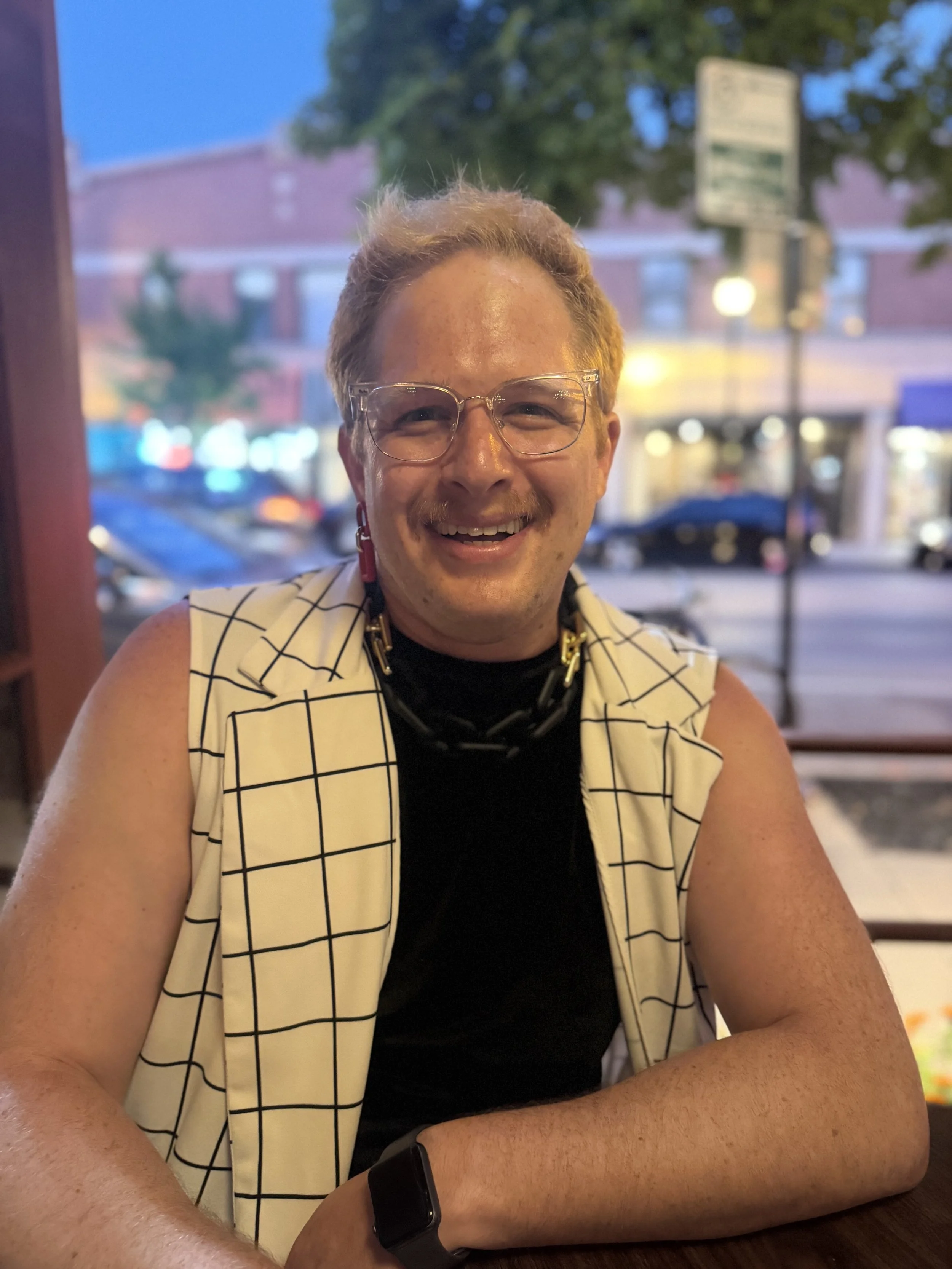 A smiling person with short, light-colored hair, wearing glasses, a black shirt, and a sleeveless checked vest, sitting at a table in a restaurant or cafe during the evening. There are trees and a building with lights in the background.