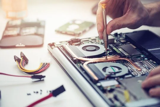 Person assembling a laptop with a screwdriver, surrounded by computer parts and hard drives on a white work surface.