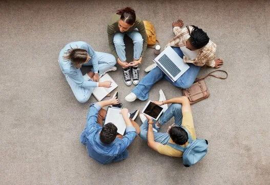 Group of five young adults sitting on the floor, using electronic devices including laptops and tablets, viewed from above.