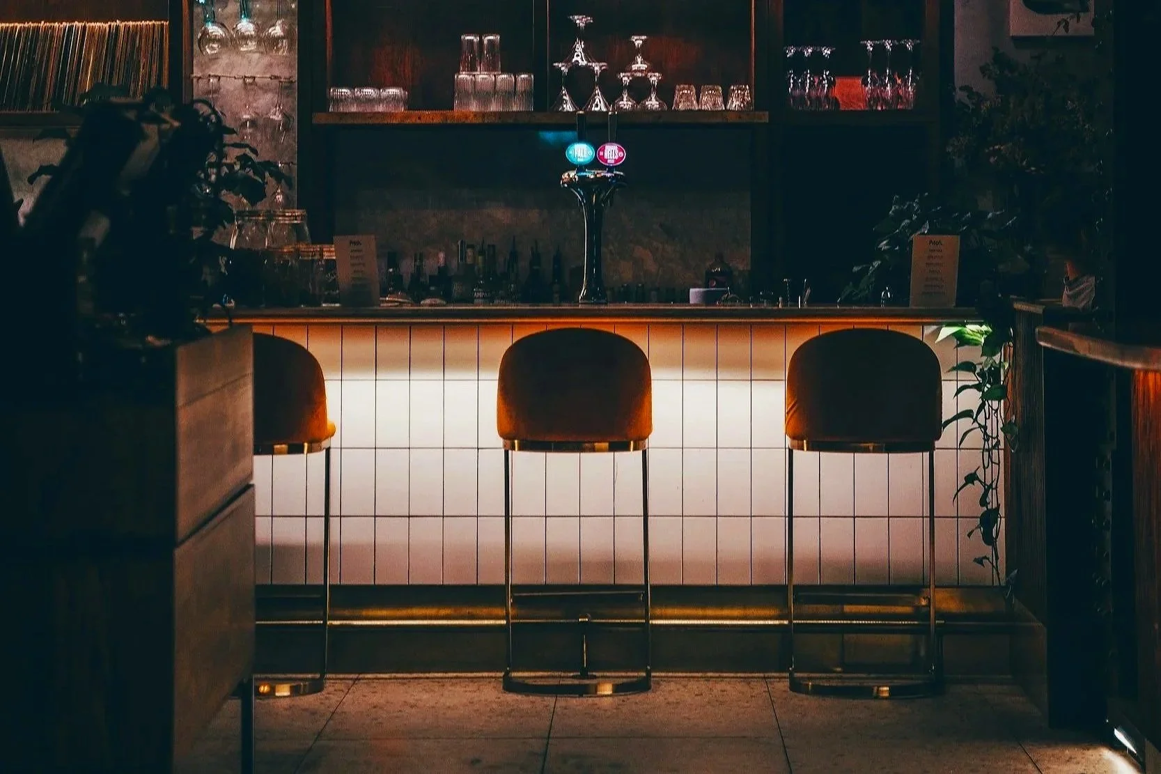 A dimly lit bar with three orange bar stools in front of a white tiled bar counter. Behind the bar, there are shelves with glasses and bottles, and a beer tap with illuminated logos.
