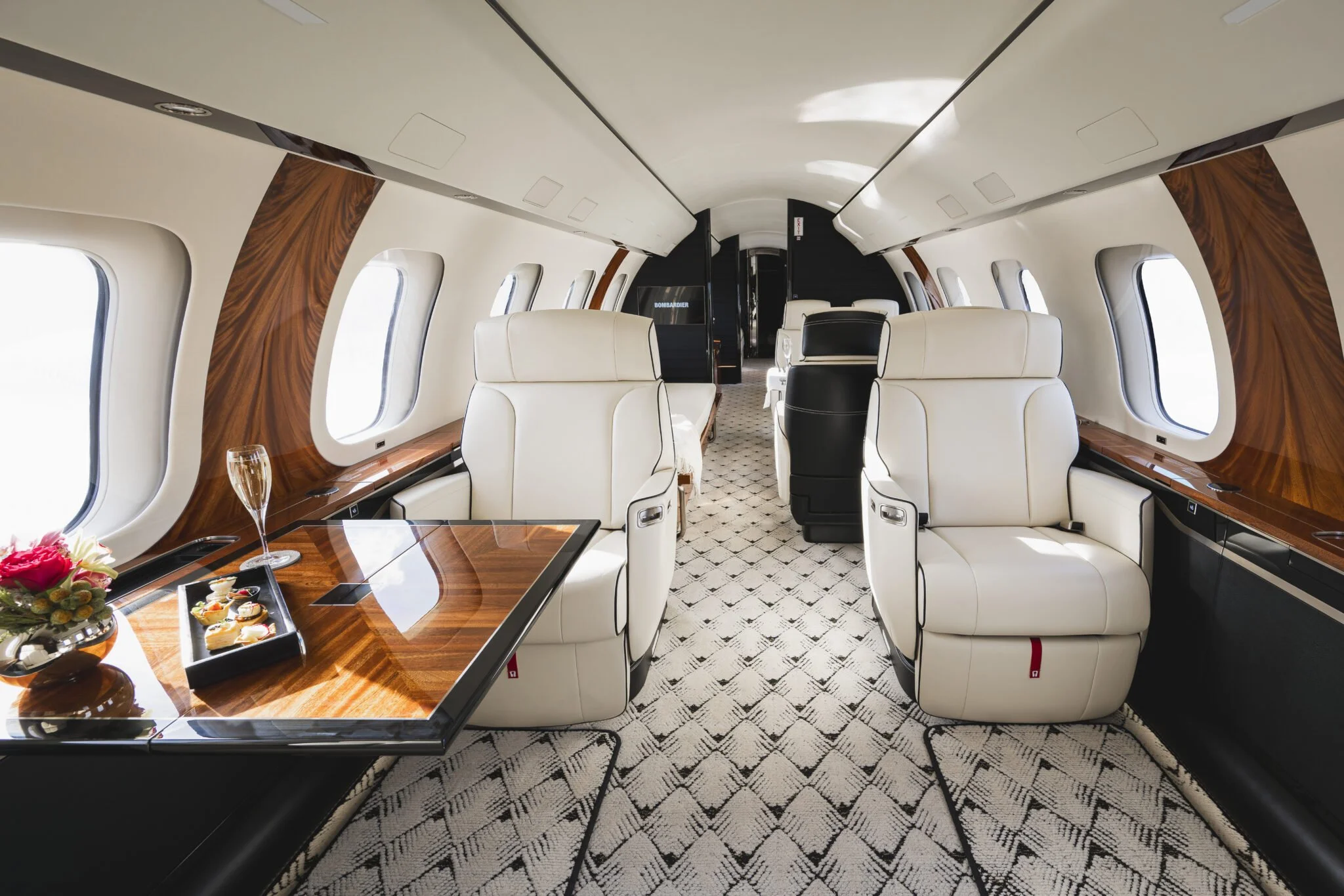 Luxury airplane cabin interior with cream-colored leather seats, wooden accents, windows along both sides, and a patterned carpet floor. There are wine glasses and a plate of desserts on the table near the front.