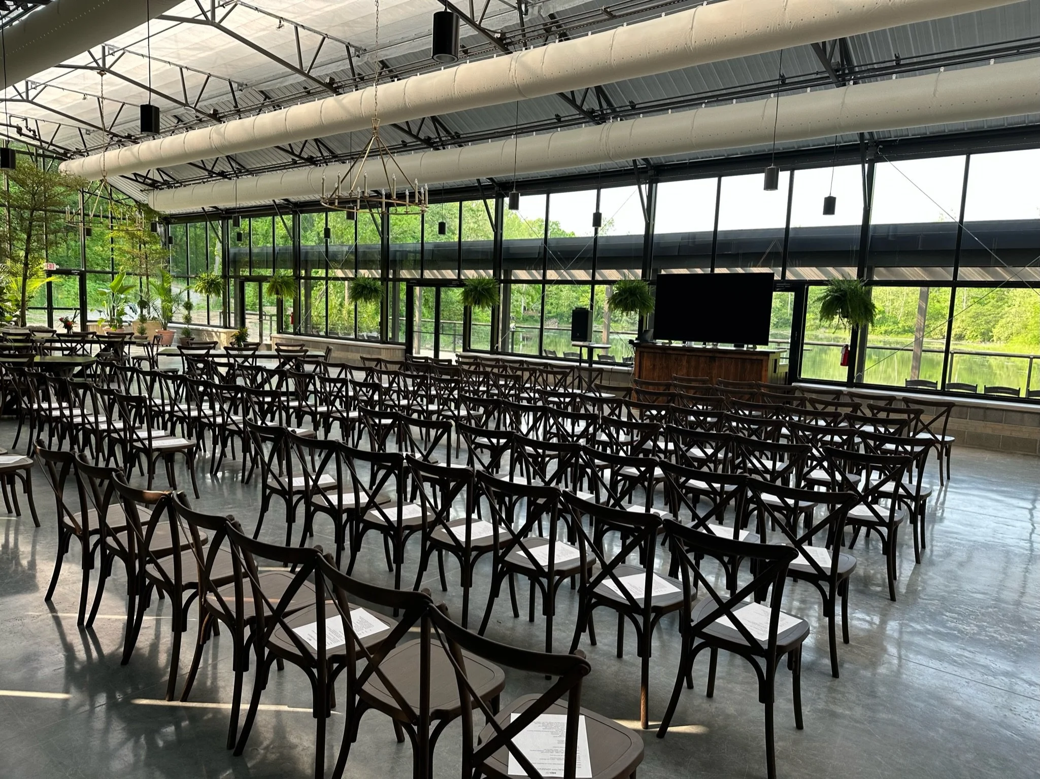Interior of The Conservatory at Evergreen, bright, airy, all-glass space with rows of chairs set up for a corporate event, hanging chandeliers, and lush green trees visible outside.