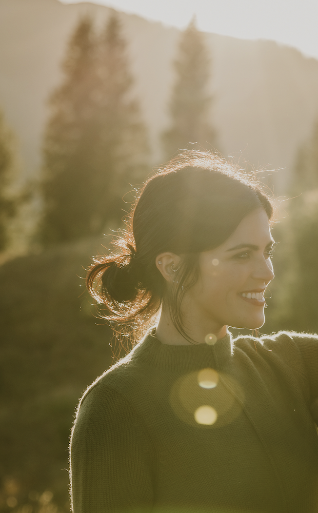 A woman with dark hair in a ponytail smiling outdoors during sunset, with trees and hills in the background.