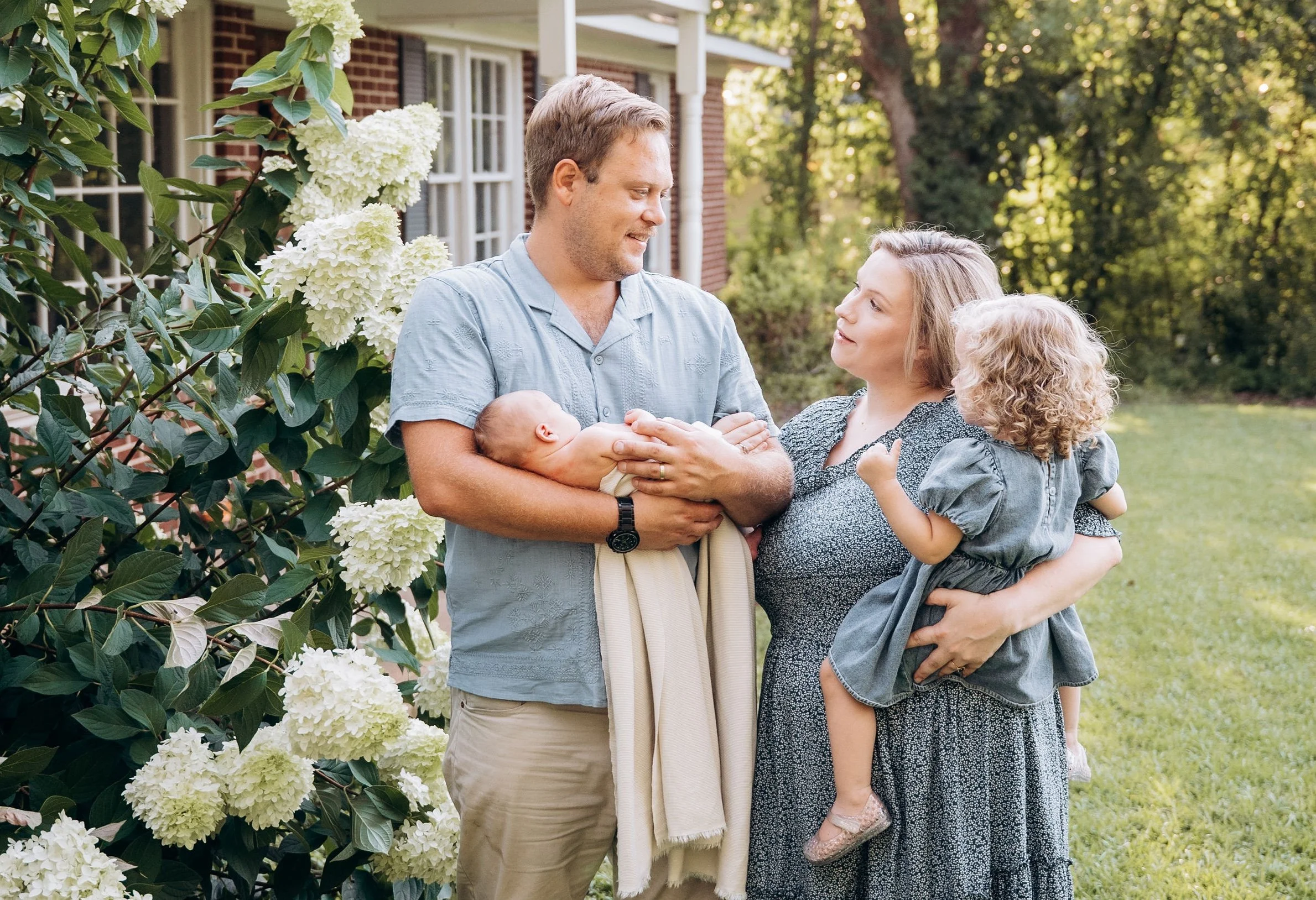 A family of four standing outside near a bush with white flowers. The father, holding a baby, and the mother, holding their young daughter, are smiling and looking at each other with a house and trees in the background.