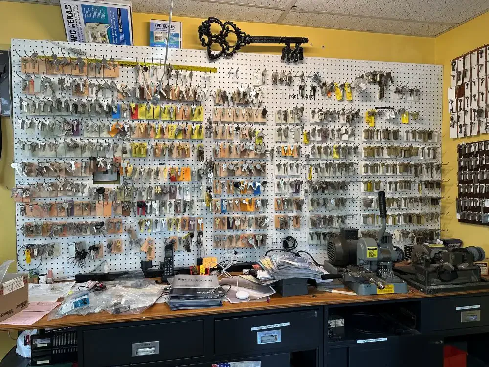 A pegboard wall with numerous keys hanging on it in a locksmith store. A large decorative key is mounted at the top. There is a workbench below with various tools, papers, and equipment, including a key-making machine.