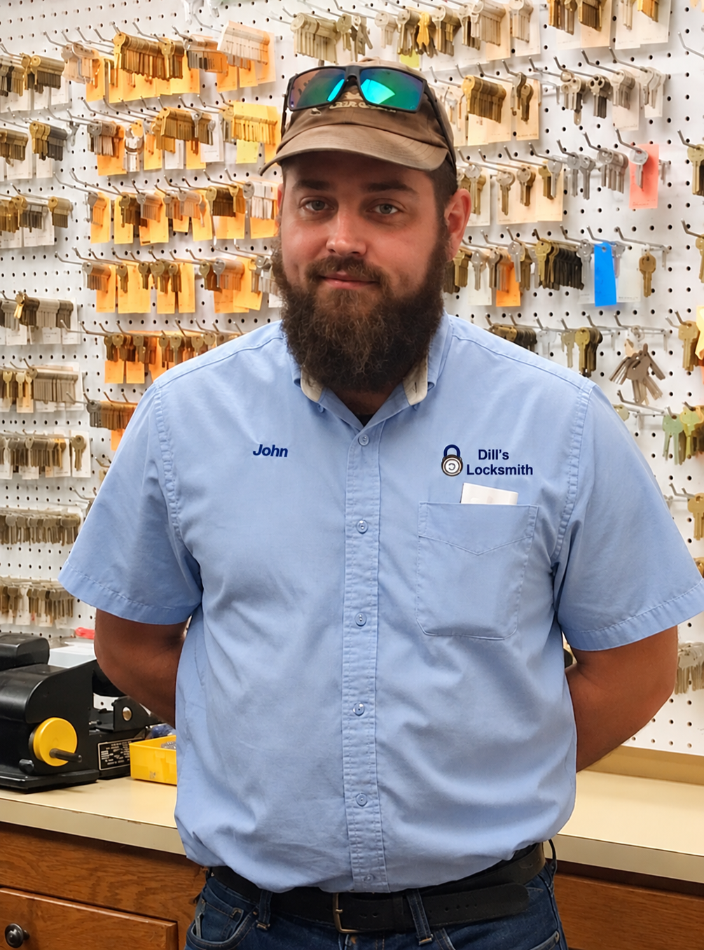 A man with a beard wearing a blue work shirt with the words 'John' and 'Dill's Locksmith' embroidered on it, standing in front of a wall of keys and key tags at a locksmith shop.