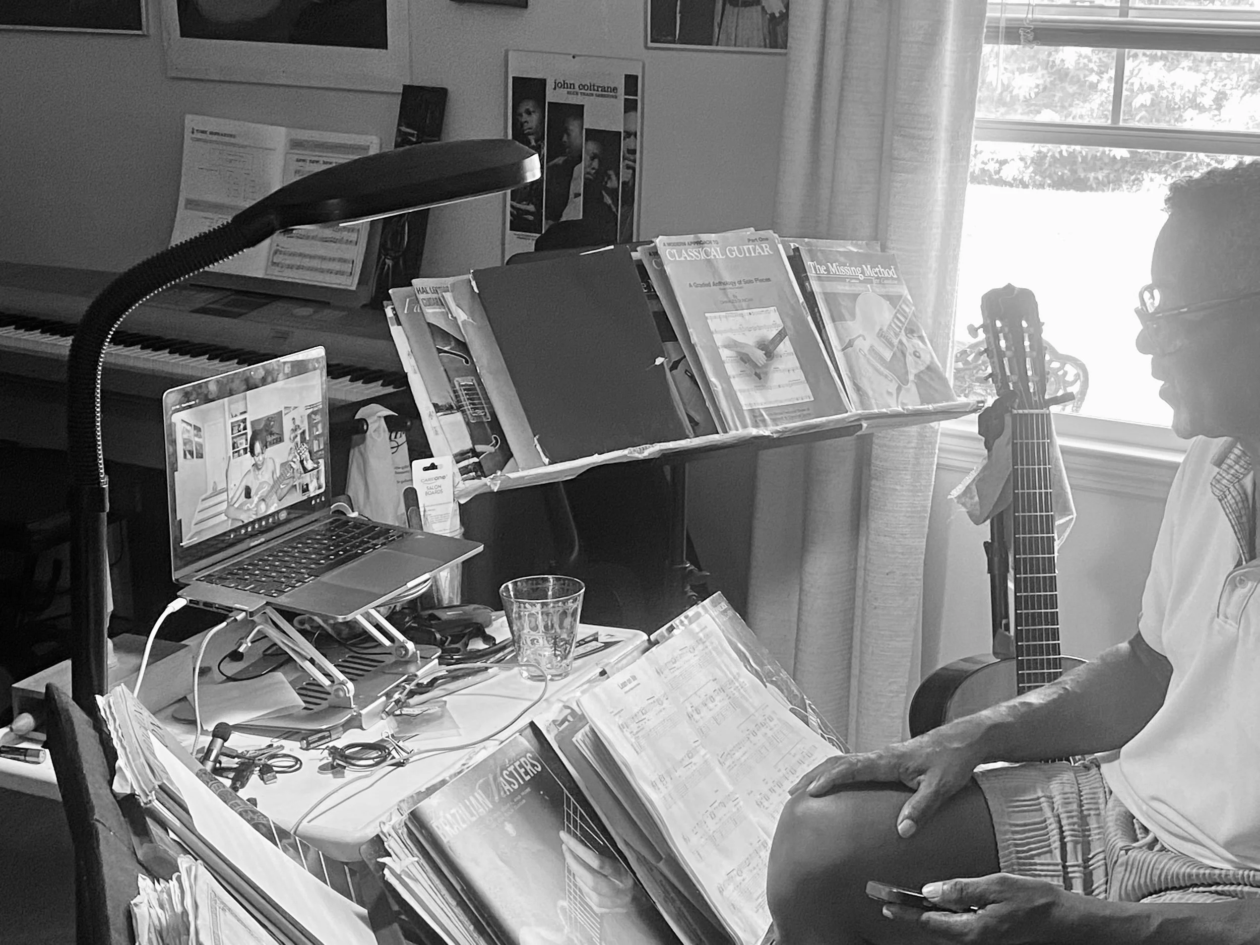 A person sitting at a cluttered desk with a guitar in hand, looking at music sheets. There are books, magazines, a laptop, a desk lamp, and posters on the wall behind them, with sunlight coming through a window.