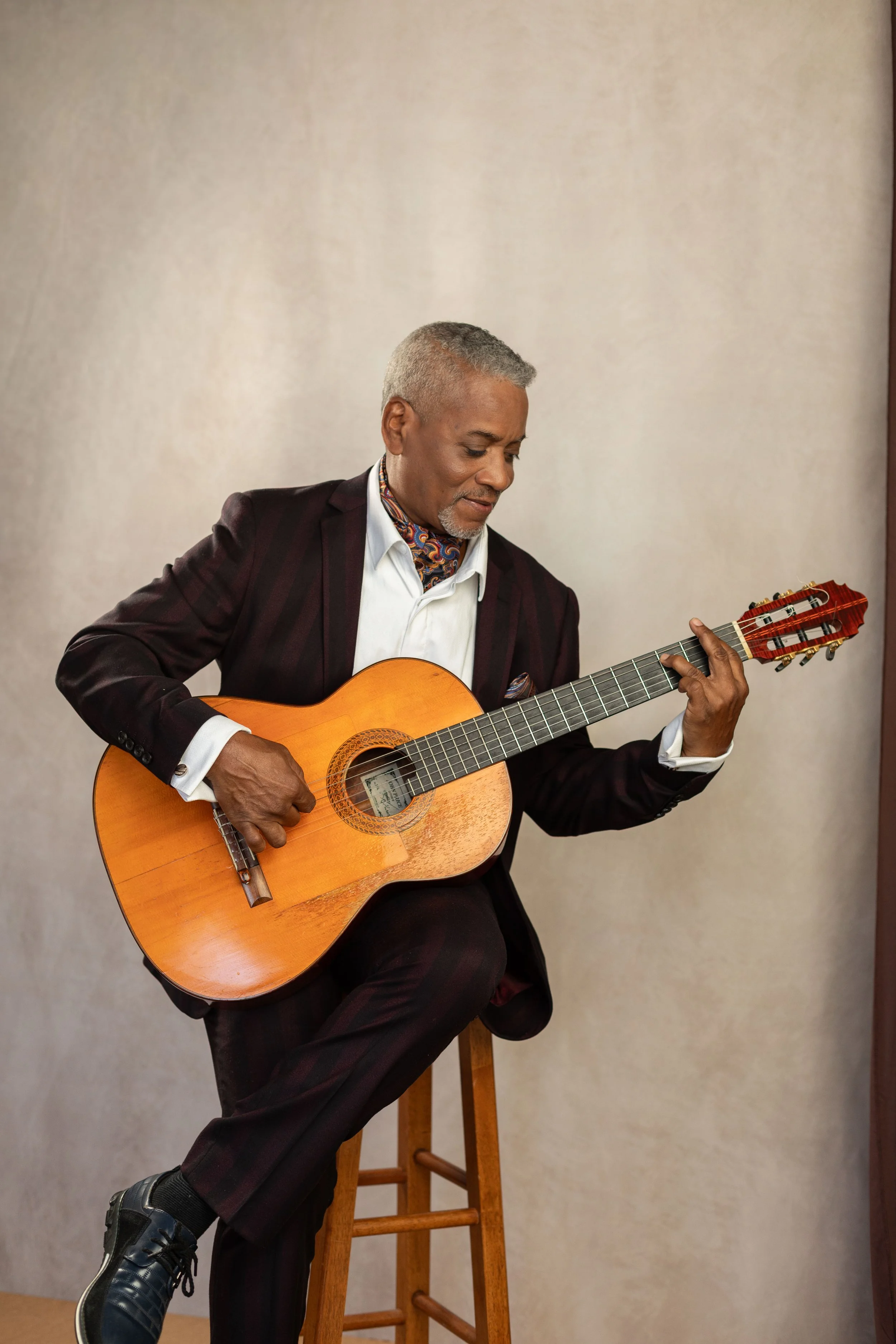 A man with salt-and-pepper hair playing an acoustic guitar while sitting on a wooden stool against a plain beige background. He is dressed in a dark suit with a colorful scarf, white shirt, and black shoes.