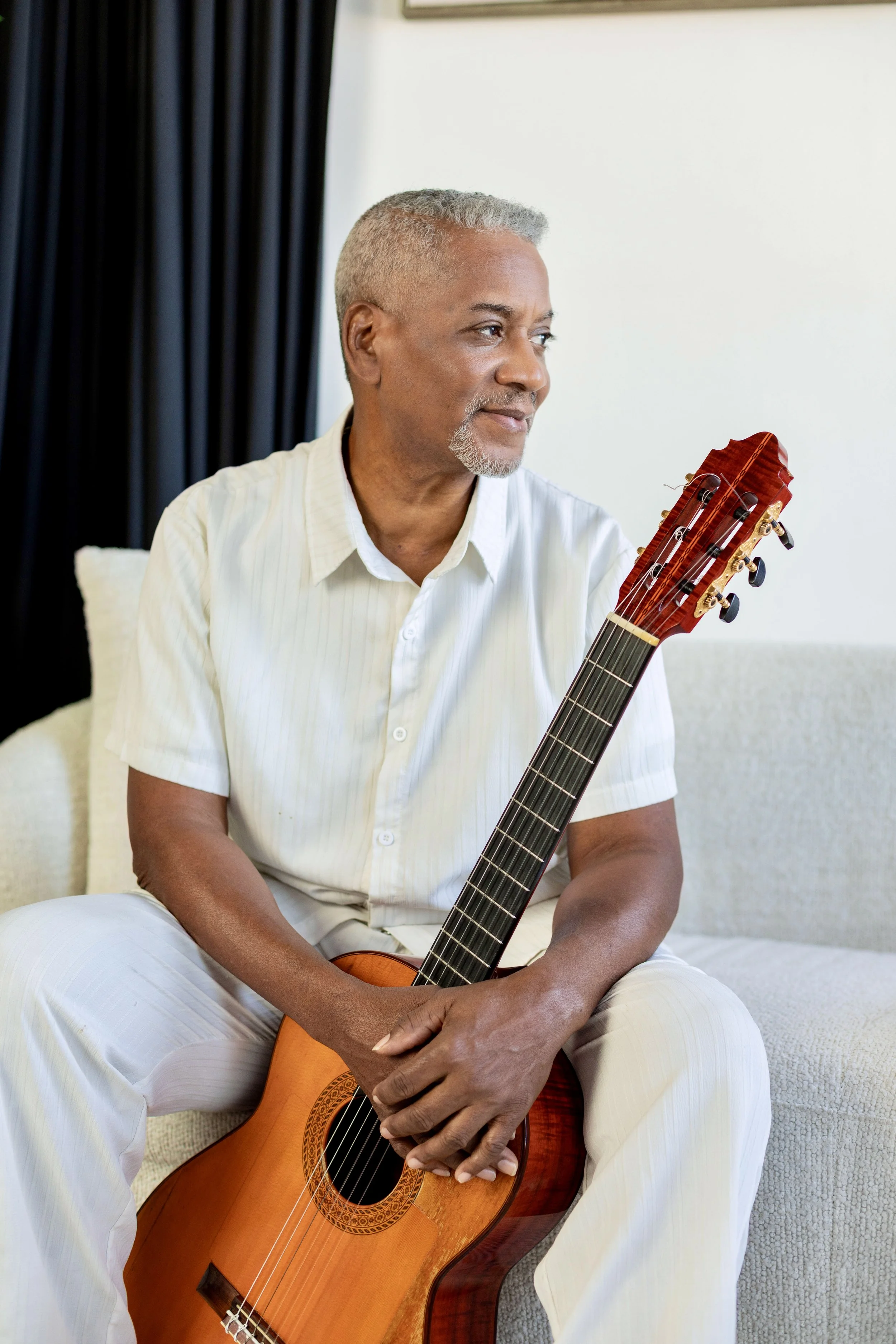 A man with short gray hair and a beard sitting on a sofa, holding an acoustic guitar, looking to his right, dressed in a white short-sleeve shirt and white pants, in a room with white walls and dark curtains.