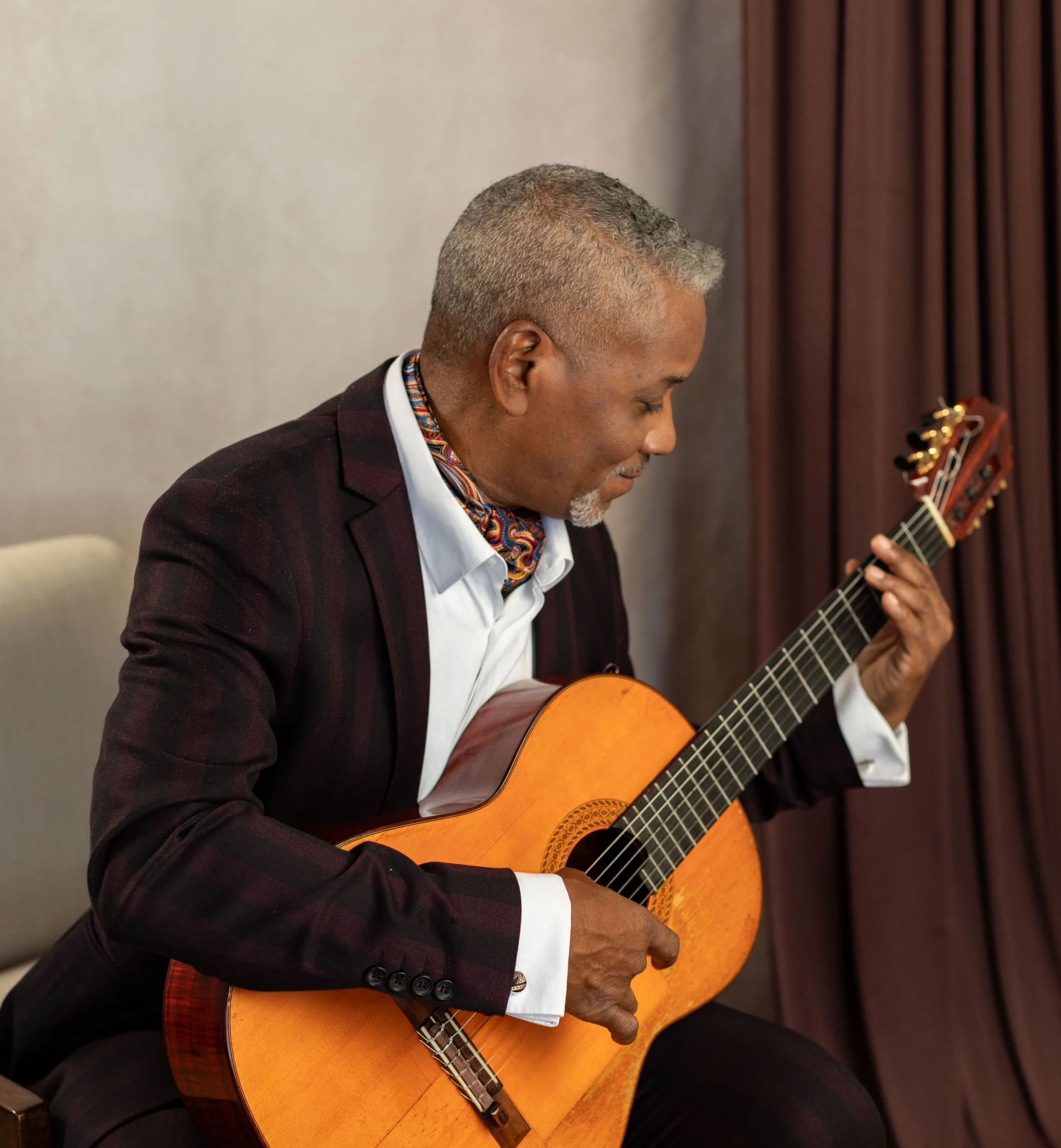 An elderly man with gray hair and a beard, dressed in a dark plaid blazer and a colorful scarf, is playing an acoustic guitar indoors in front of a beige wall and brown curtains.